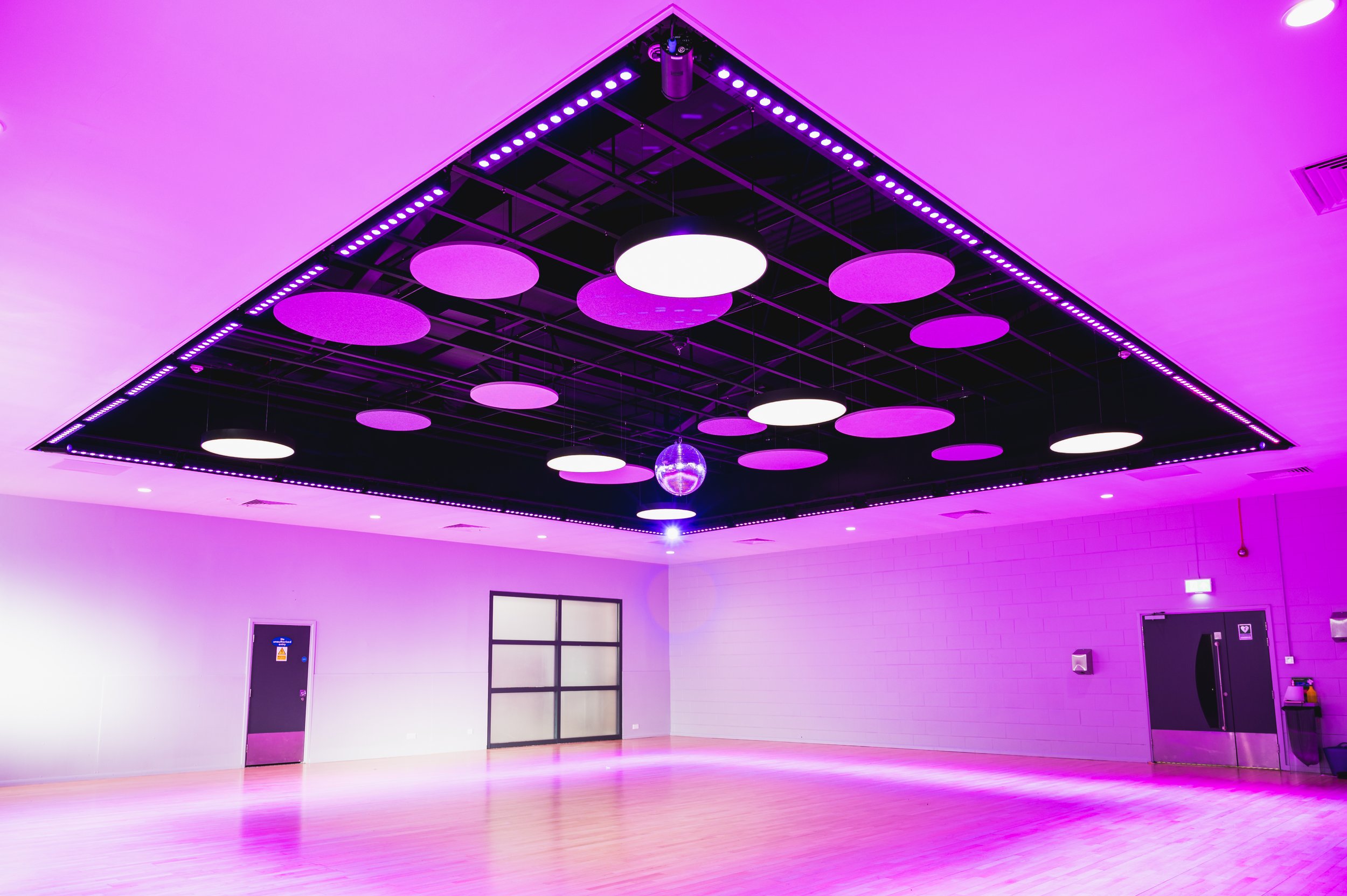 A dance studio with a black ceiling grid, purple and white circular ceiling lights, purple LED strip lights, a light-colored wooden floor, white brick wall, door, and window.