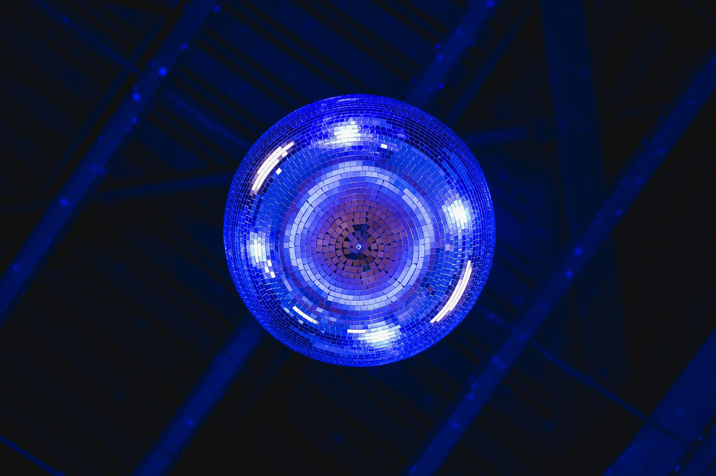 A blue disco ball hanging from a ceiling with visible metal beams, reflecting light in a dark setting.