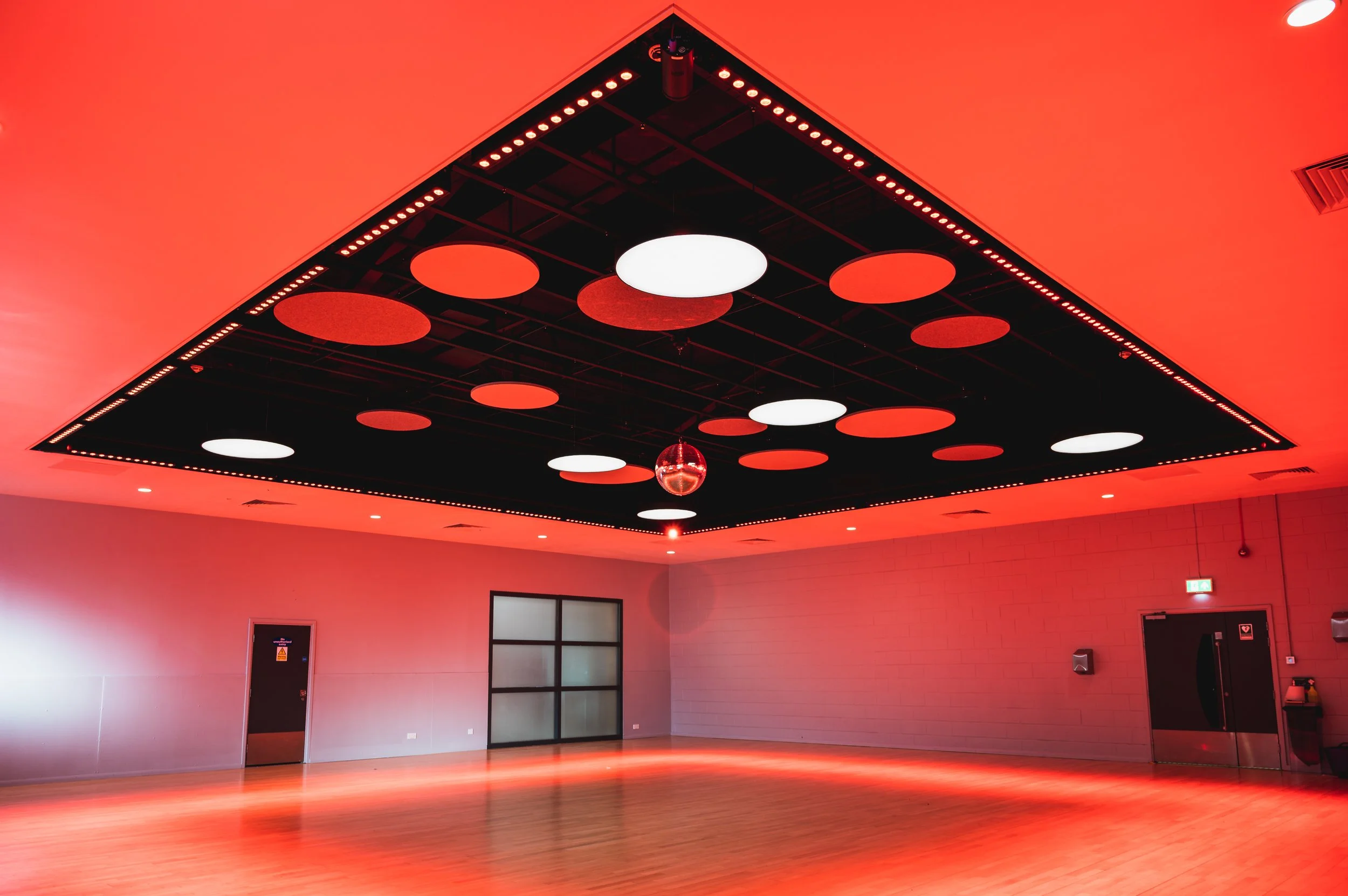 Empty dance or event room with a wooden floor, beige brick walls, a black and red ceiling with round red and white lights, and a disco ball hanging from the ceiling.