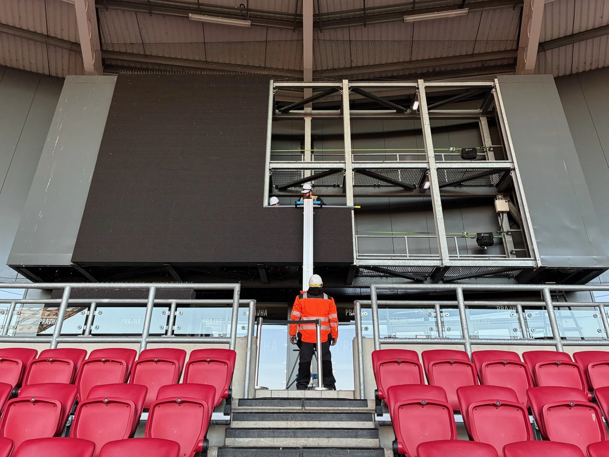 Workers installing a large LED screen in a stadium with red seats in the foreground.