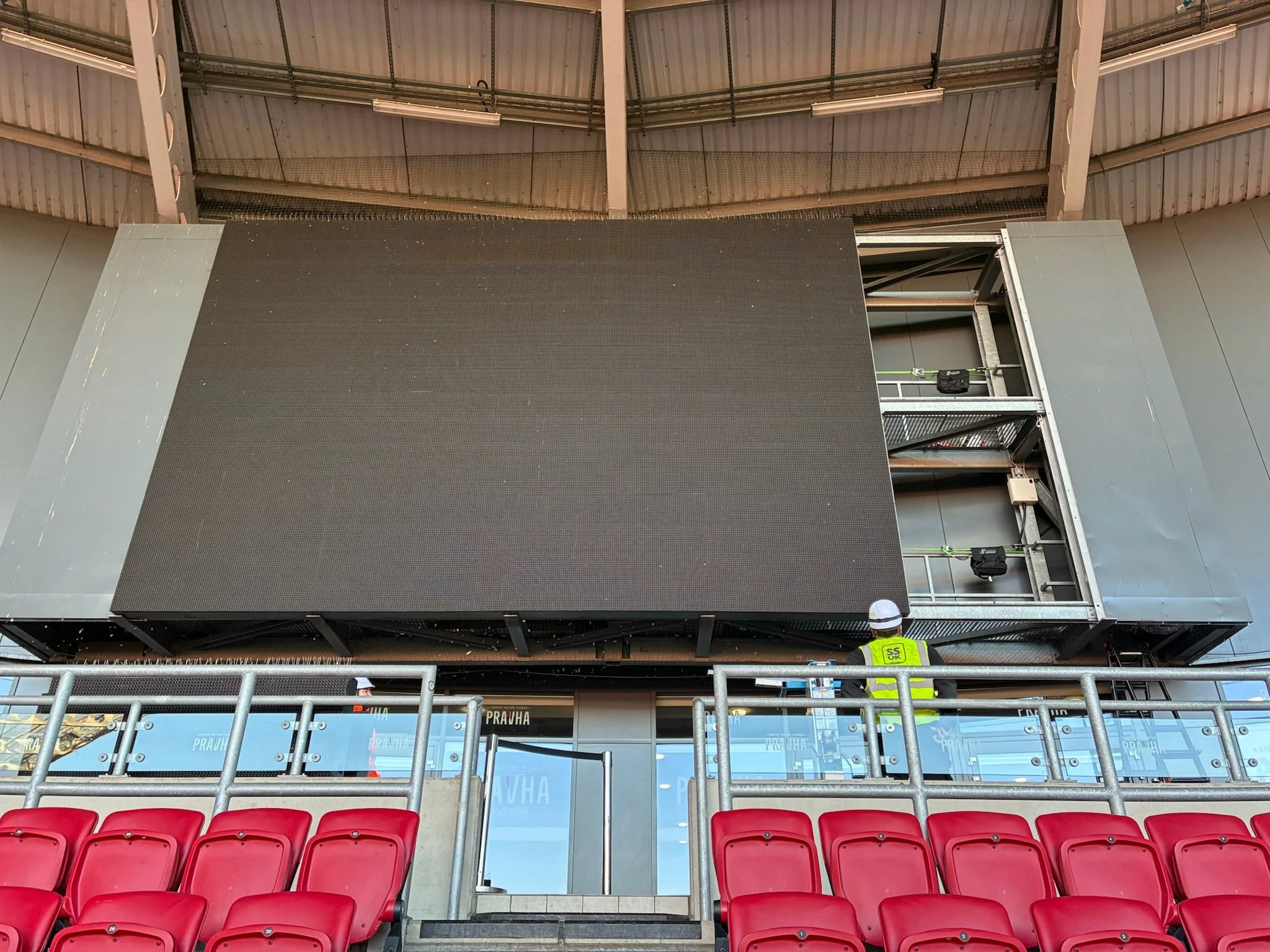 Inside a stadium with red seats in the foreground, construction workers wearing safety helmets and vests working on a large screen or wall on the upper level, with the roof structure visible above.