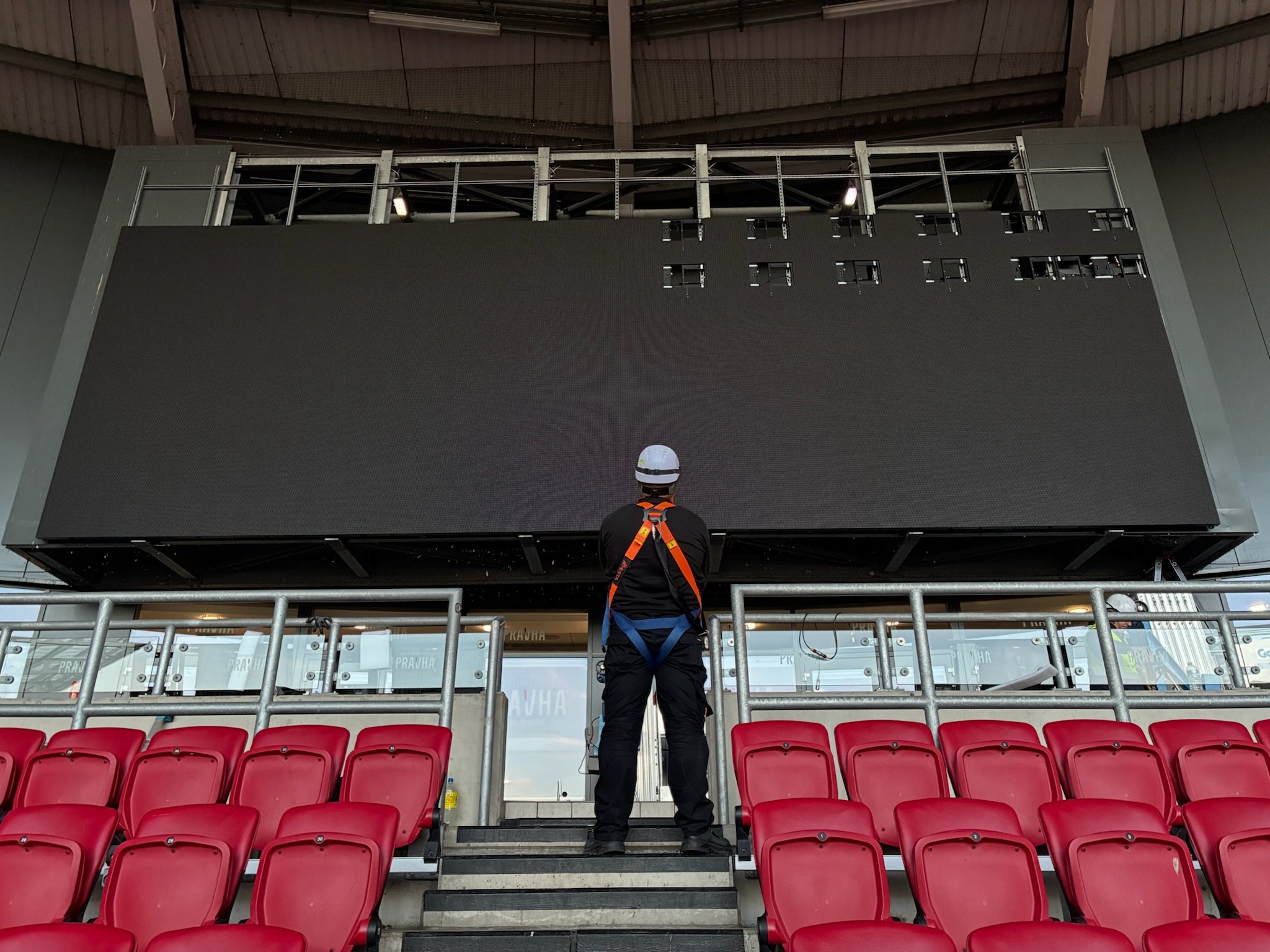 A man wearing a helmet and a harness is installing or maintaining a large LED screen in a sports stadium or arena, with red seats in the foreground and an unfinished upper section of the screen.