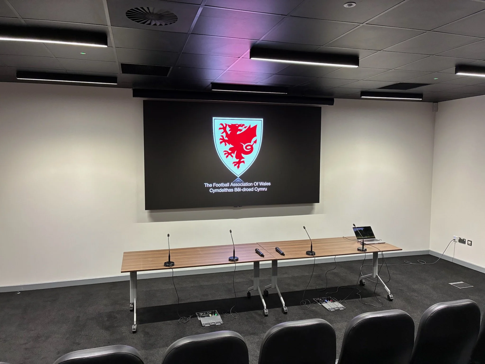 A conference room with a large screen displaying the logo of the Football Association of Wales and Welsh text. The room has a long table with microphones, laptops, and chairs in front of it. The ceiling has modern black panel lighting.