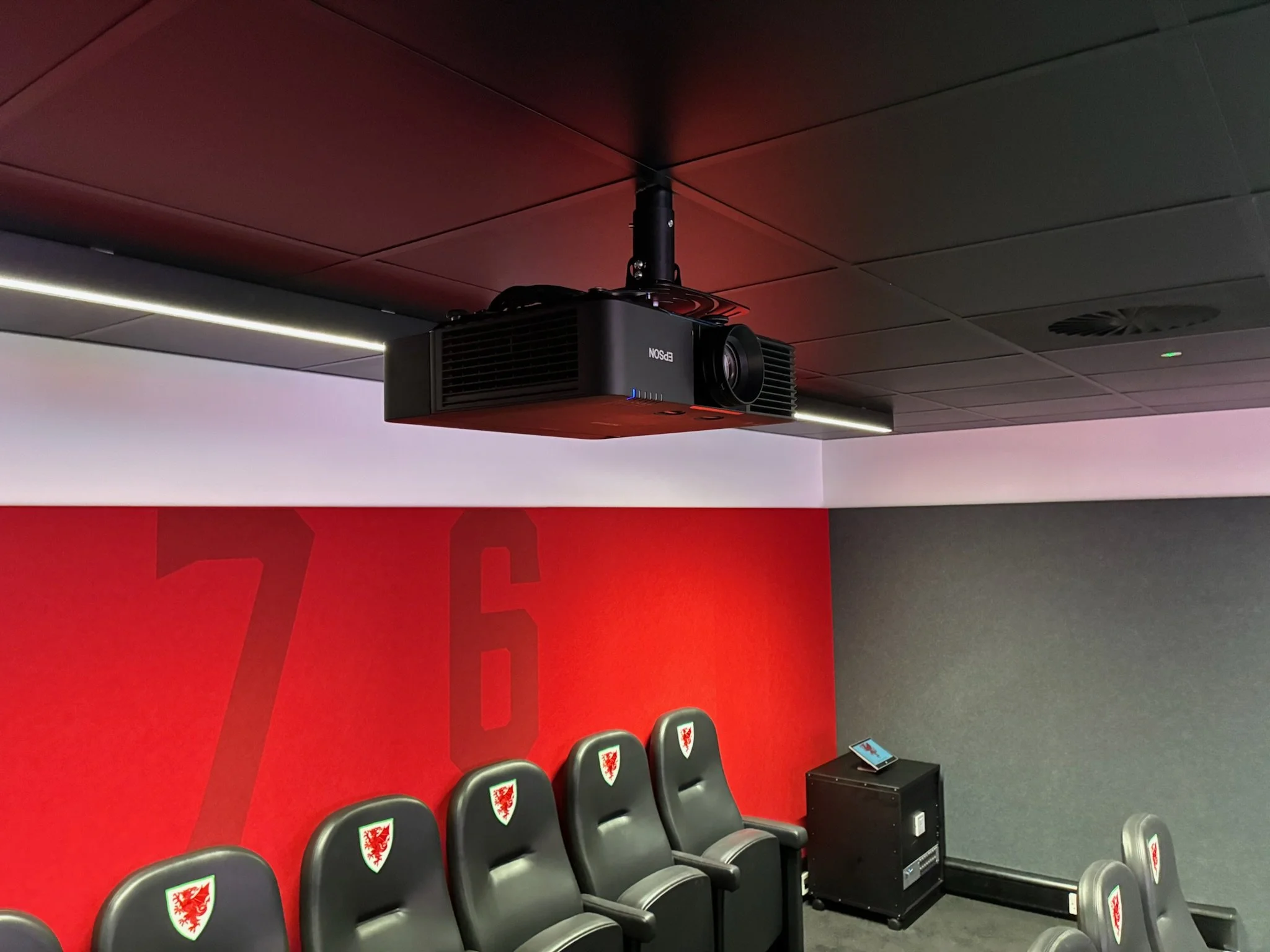 A row of black chairs with the Welsh flag shield on the headrests, placed against a red wall with a large Welsh emblem, in a room equipped with a projector mounted on the ceiling, a small black table with a phone and power strip, and part of a display screen showing a website.