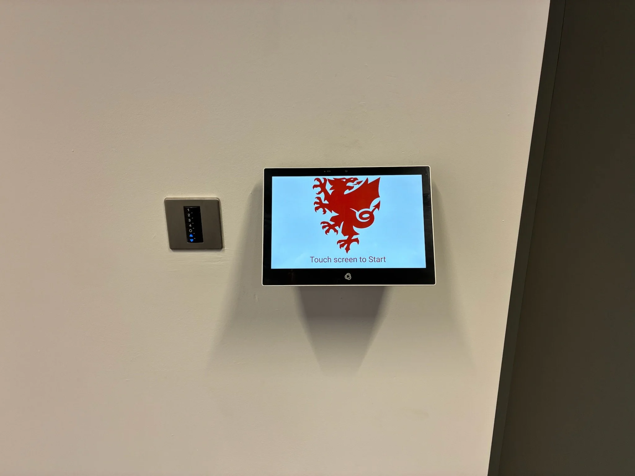 A touchscreen device on a white wall displaying a red lion graphic and the words "Touch screen to Start."