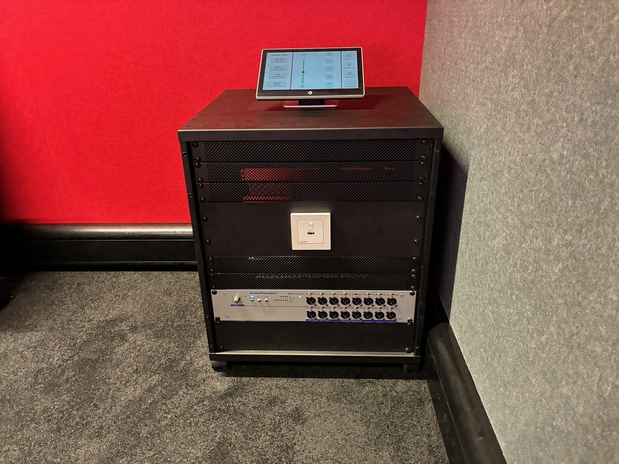 A black audio equipment rack with a control panel on top, placed against a red and gray wall, with a power outlet at the bottom, and a gray carpeted floor.