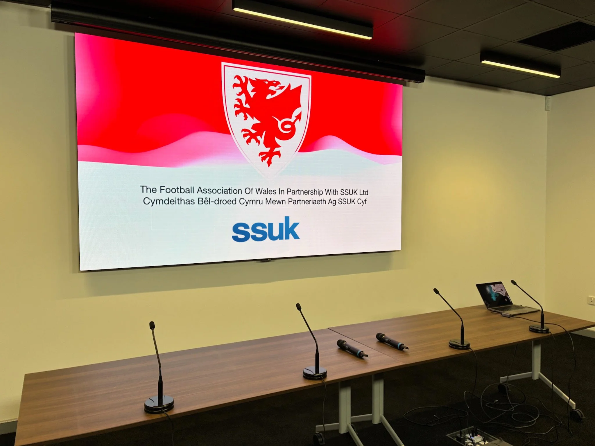 Conference room with a large screen displaying the Welsh Football Association logo and text, microphones on the table, and a laptop, set up for a presentation or meeting.