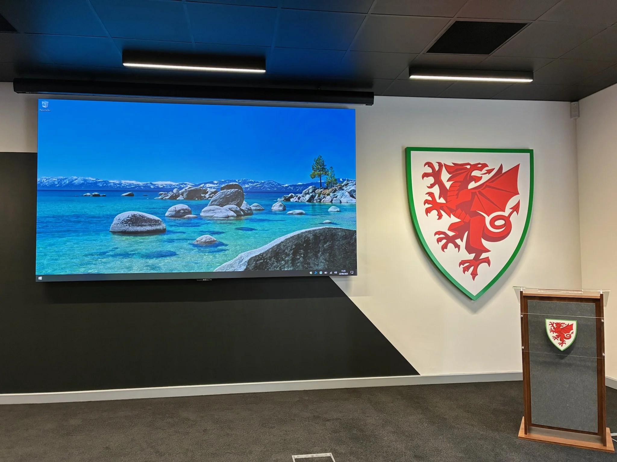 Interior of a room with a large digital screen showing a landscape of a lake with rocks, trees, and mountains in the background. To the right of the screen is a shield emblem with a red dragon on a white background outlined in green. There are ceiling lights and an additional sign with the same emblem on a stand in the foreground.