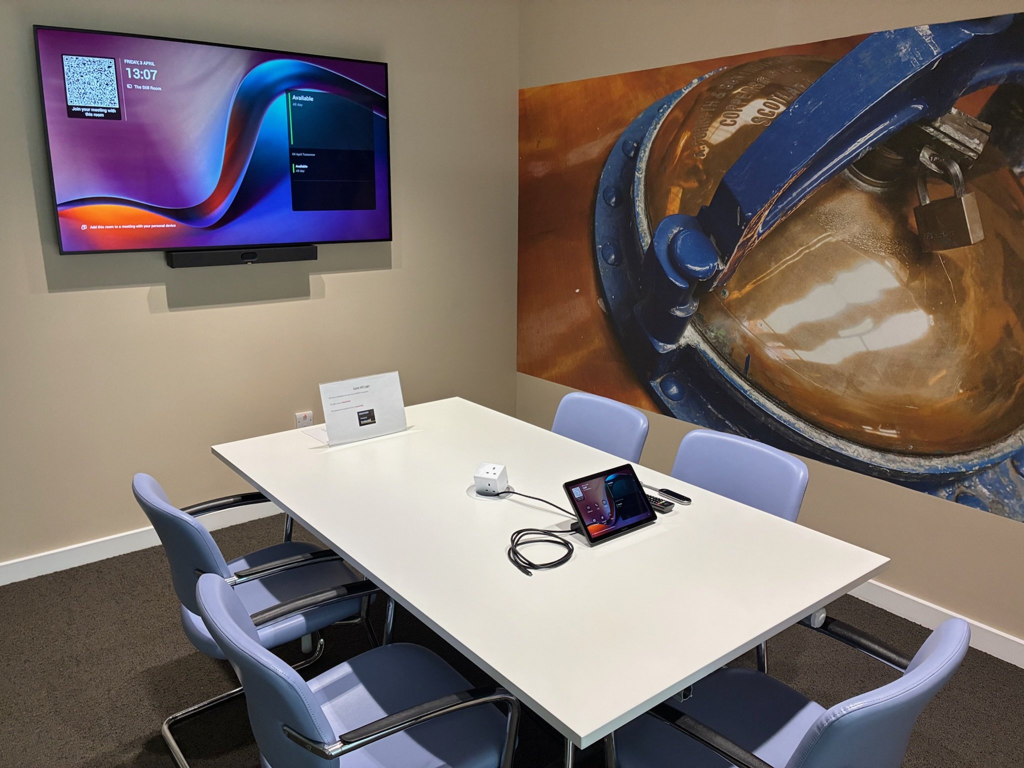 A modern conference room with a white table, six blue chairs, and electronic devices including a tablet, smartphone, and charging cables. A large wall mural of a close-up of a mechanical part is on the right wall. A flat-screen television displays a digital interface on the left wall.