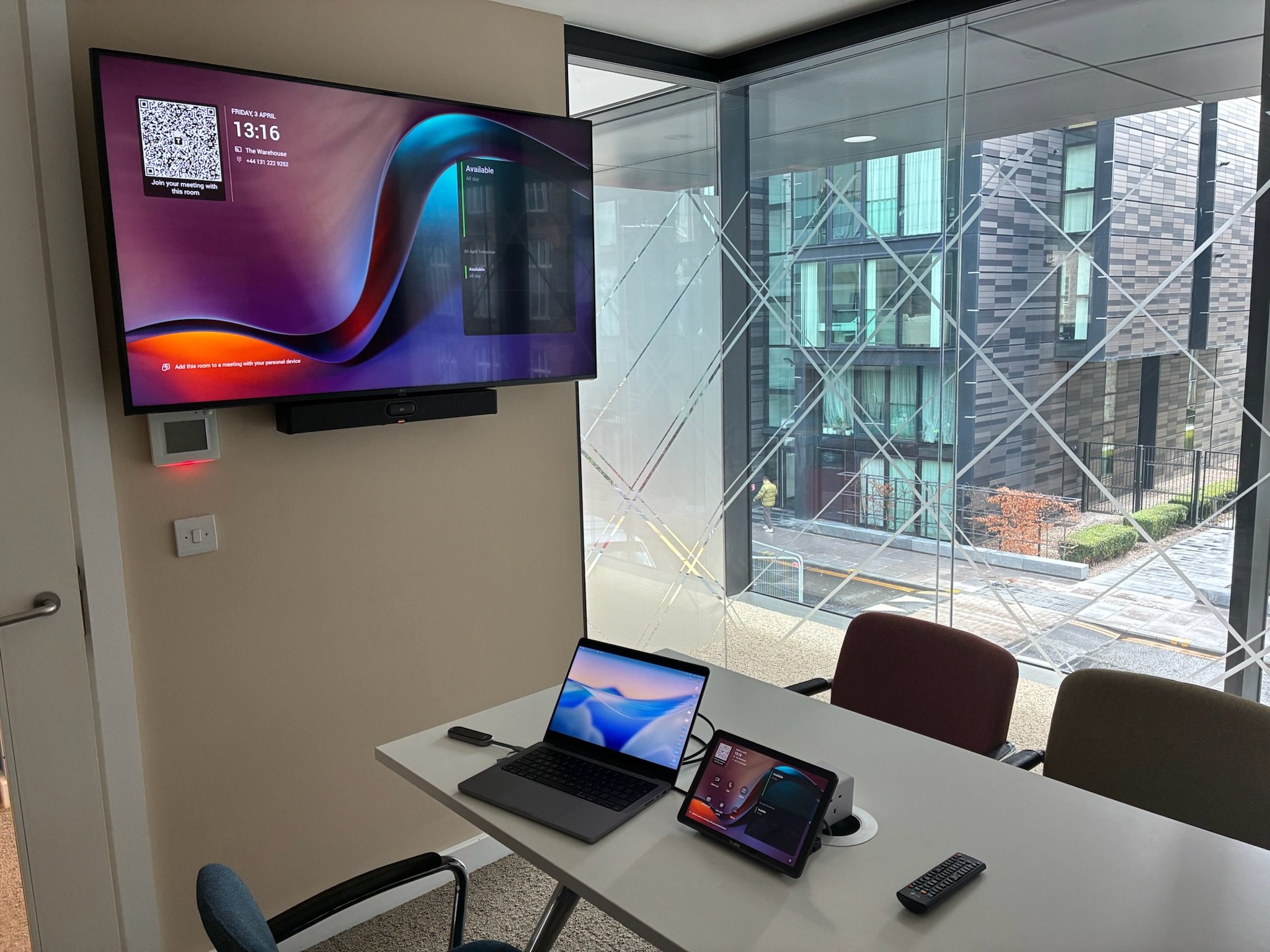 An indoor office or conference room with a large wall-mounted monitor displaying the time, date, and room information. A table with laptops and a tablet, and chairs are visible. Through the glass wall, a modern building exterior with windows and some outdoor trees can be seen.