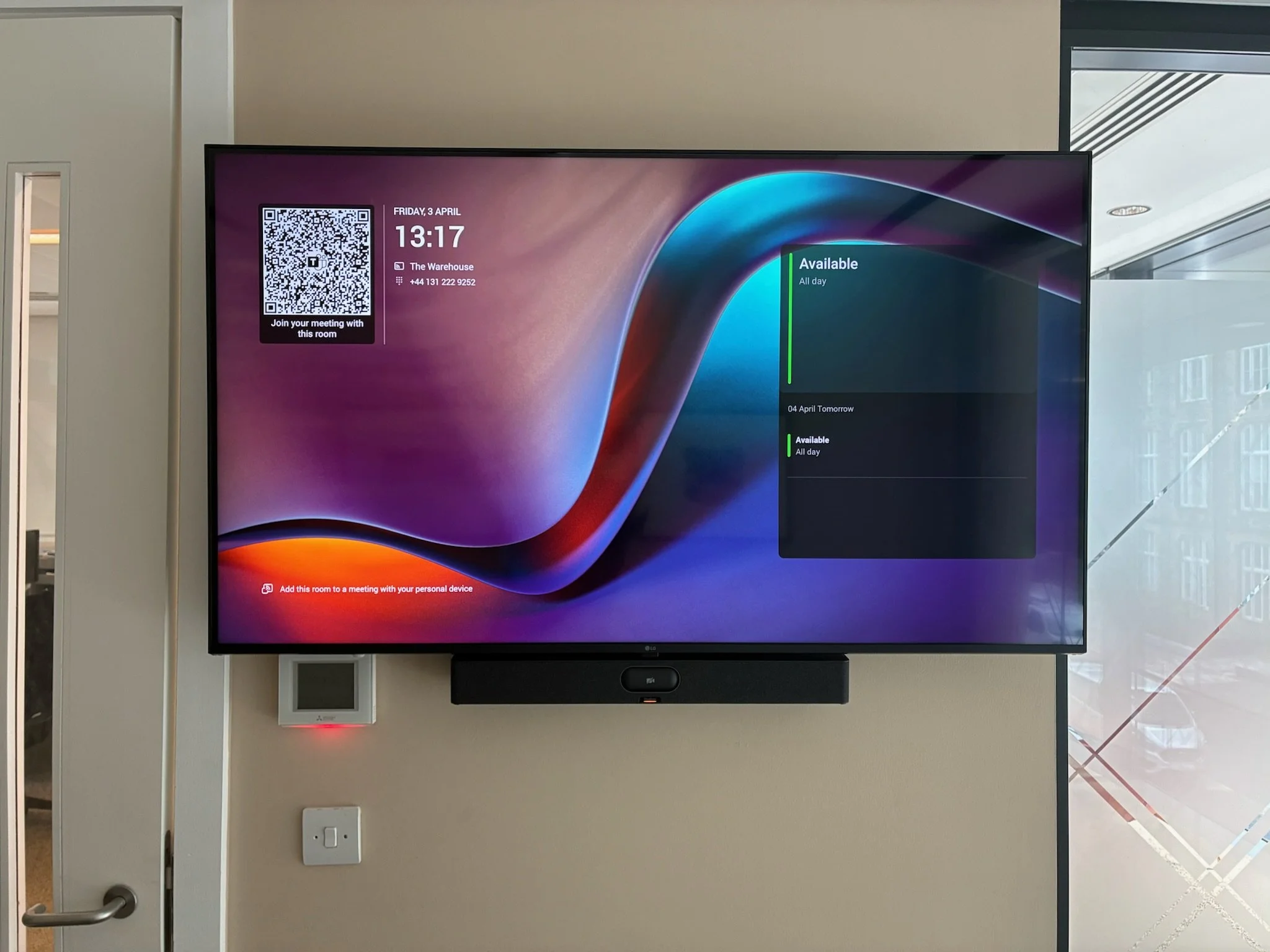 A large digital display screen showing the date, time, and meeting room information, including a QR code for joining a meeting, mounted on a beige wall in an office environment.