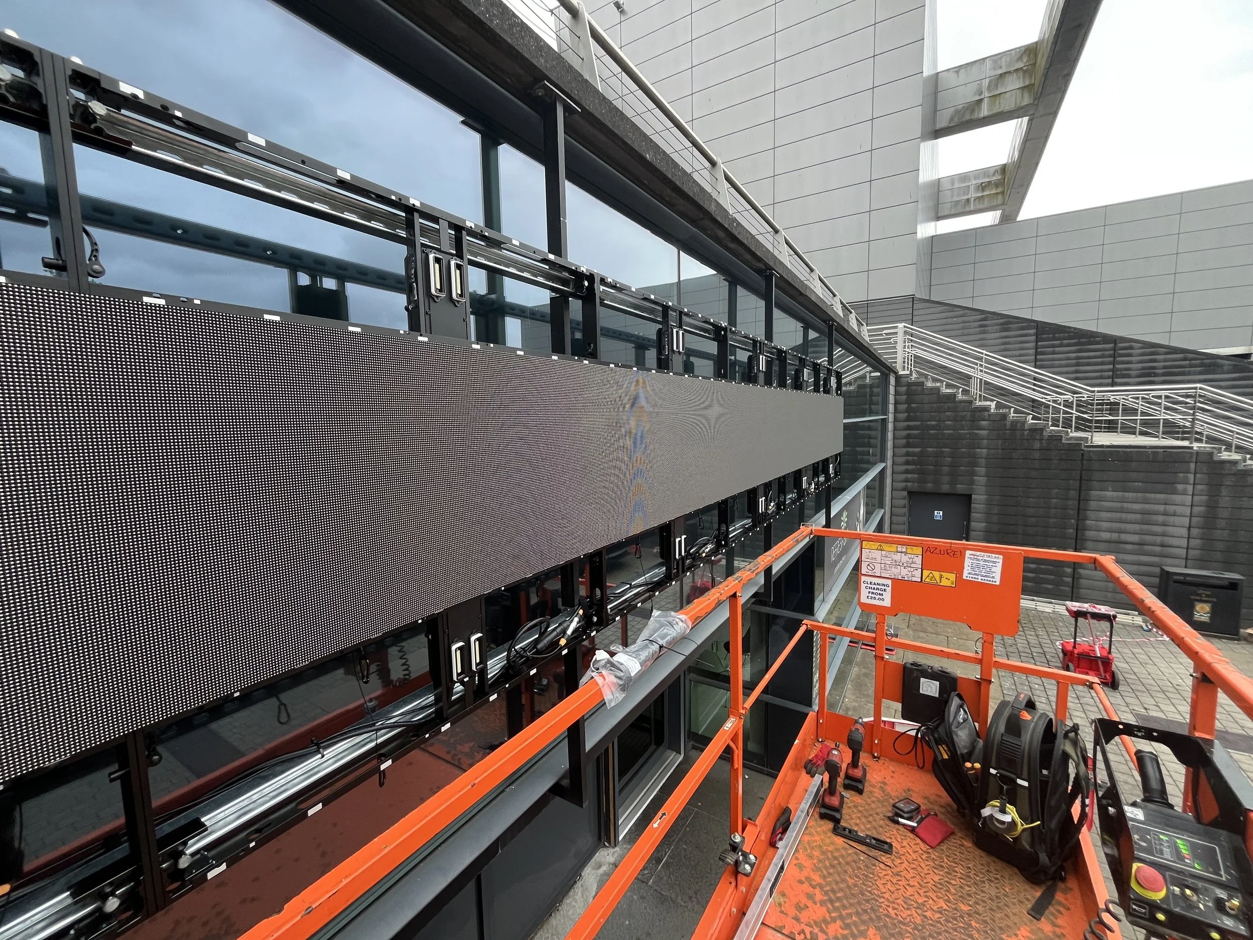 Construction or maintenance work on a modern building exterior, with progress on digital display panels, scaffolding, and construction tools and materials.