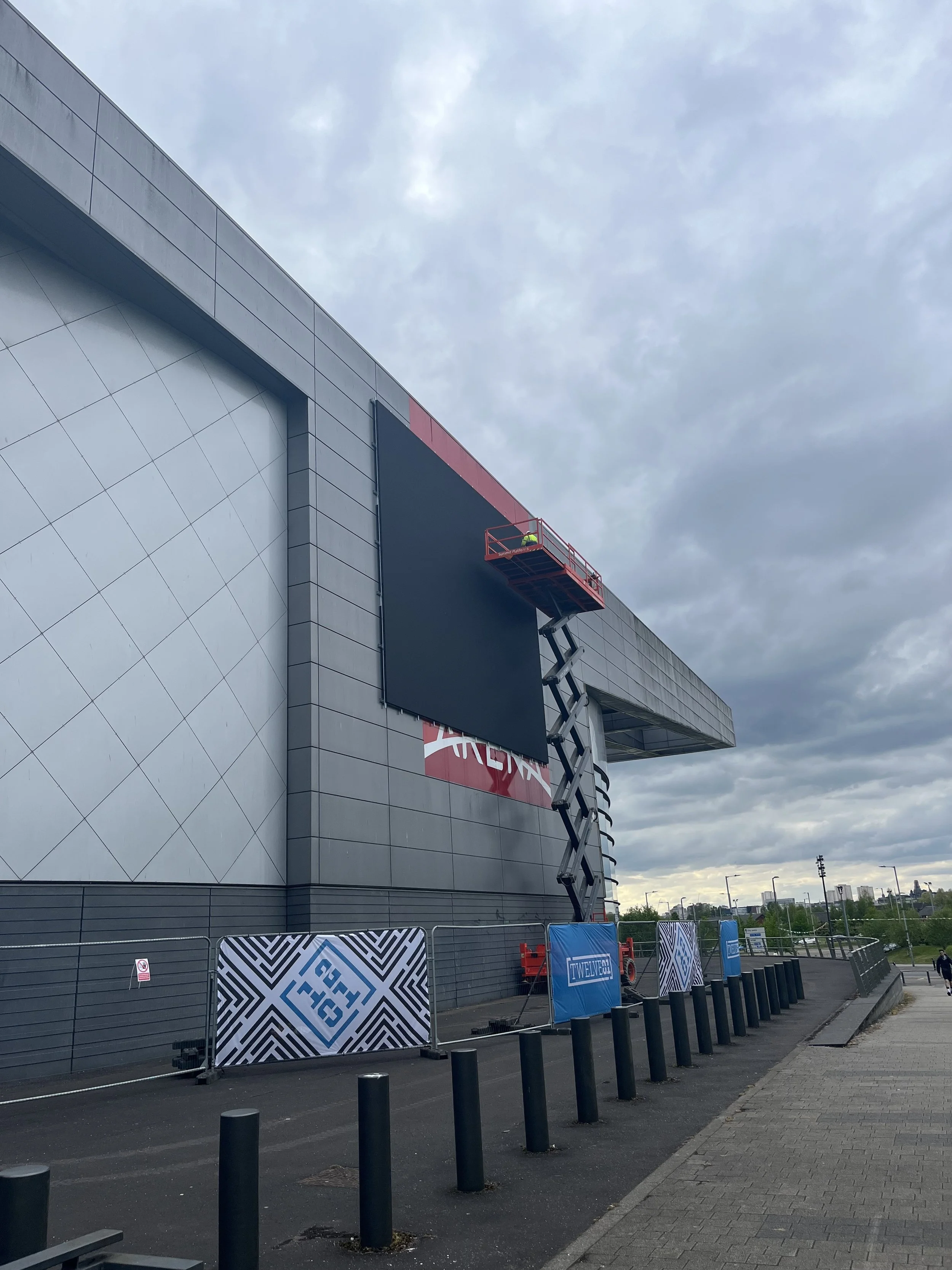 Construction scaffolding and large black screen on the exterior of a modern building with a cloudy sky above, surrounded by fencing and parking lot.