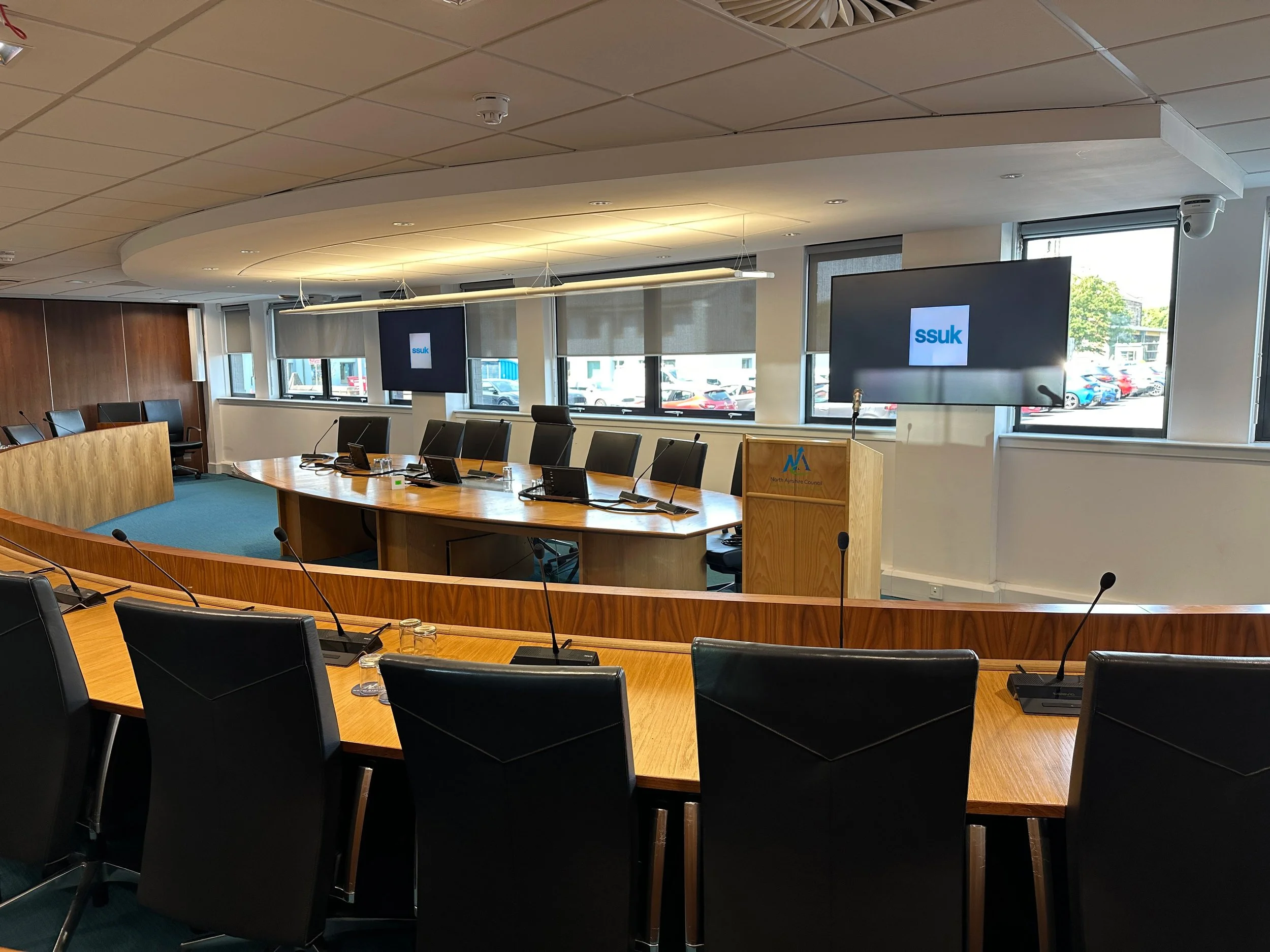 Empty conference room with a long wooden table, black chairs, multiple microphones, large windows, and two screens displaying the SSUK logo.