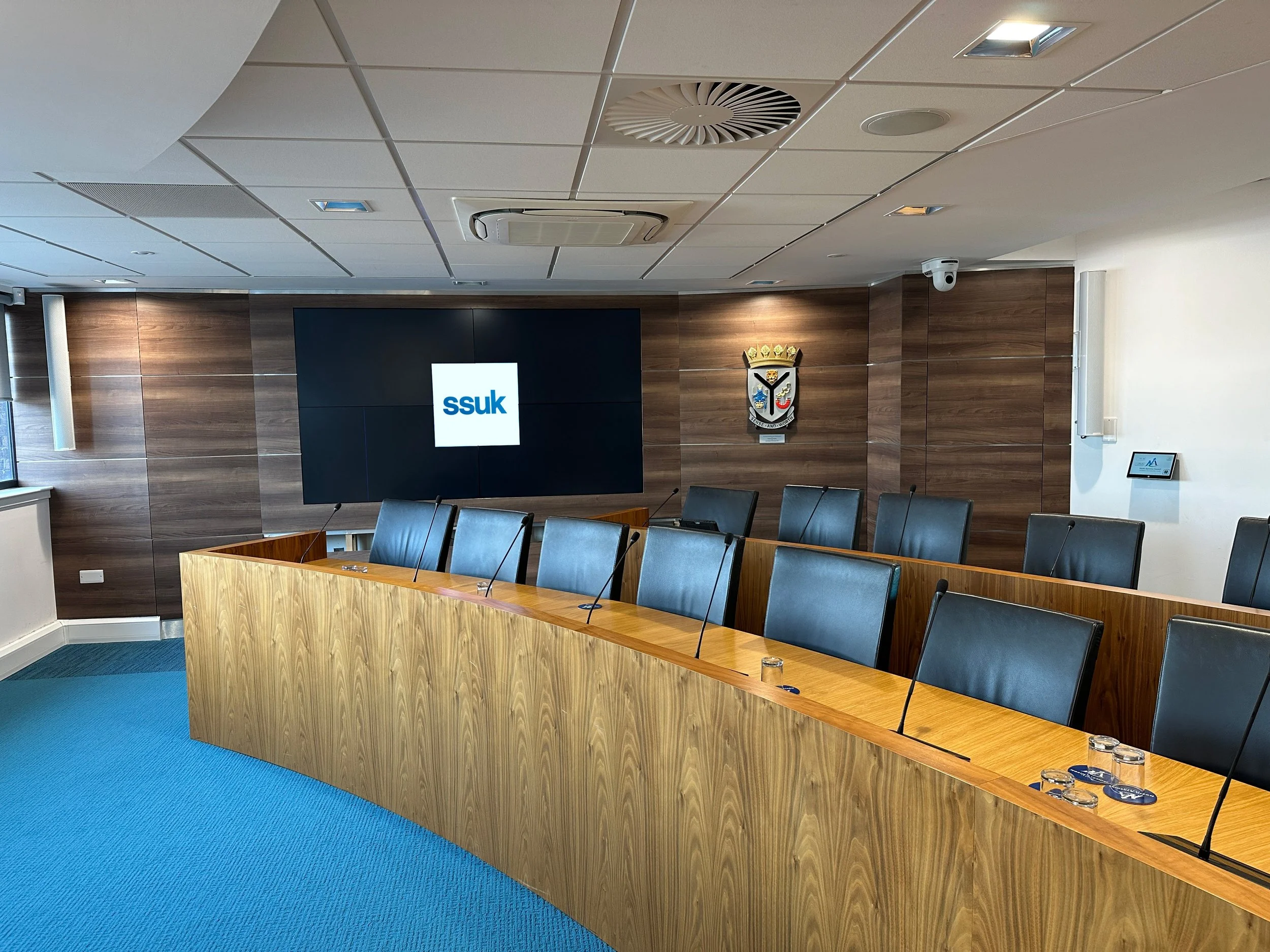 A conference room with a wooden table and black chairs, a large digital screen displaying 'ssuk,' and a crest on the wall with a crown, shield, and banners.