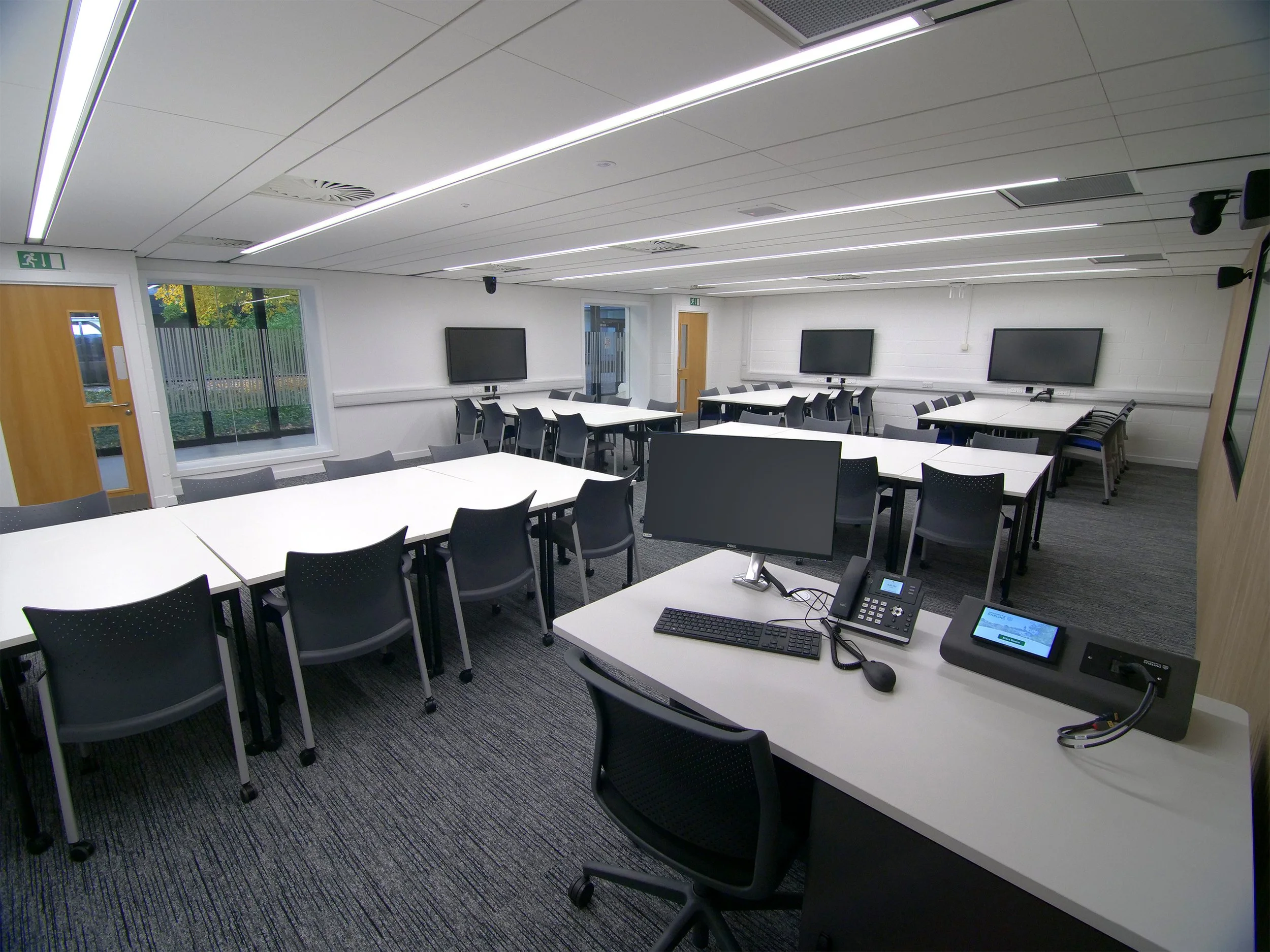 A modern classroom with white tables, black chairs, and multiple large television screens mounted on white walls. There are computers, a telephone, and a conference phone on the front desk. The ceiling has white panels with linear LED lights, and there is a glass door with an exit sign and windows showing trees outside.
