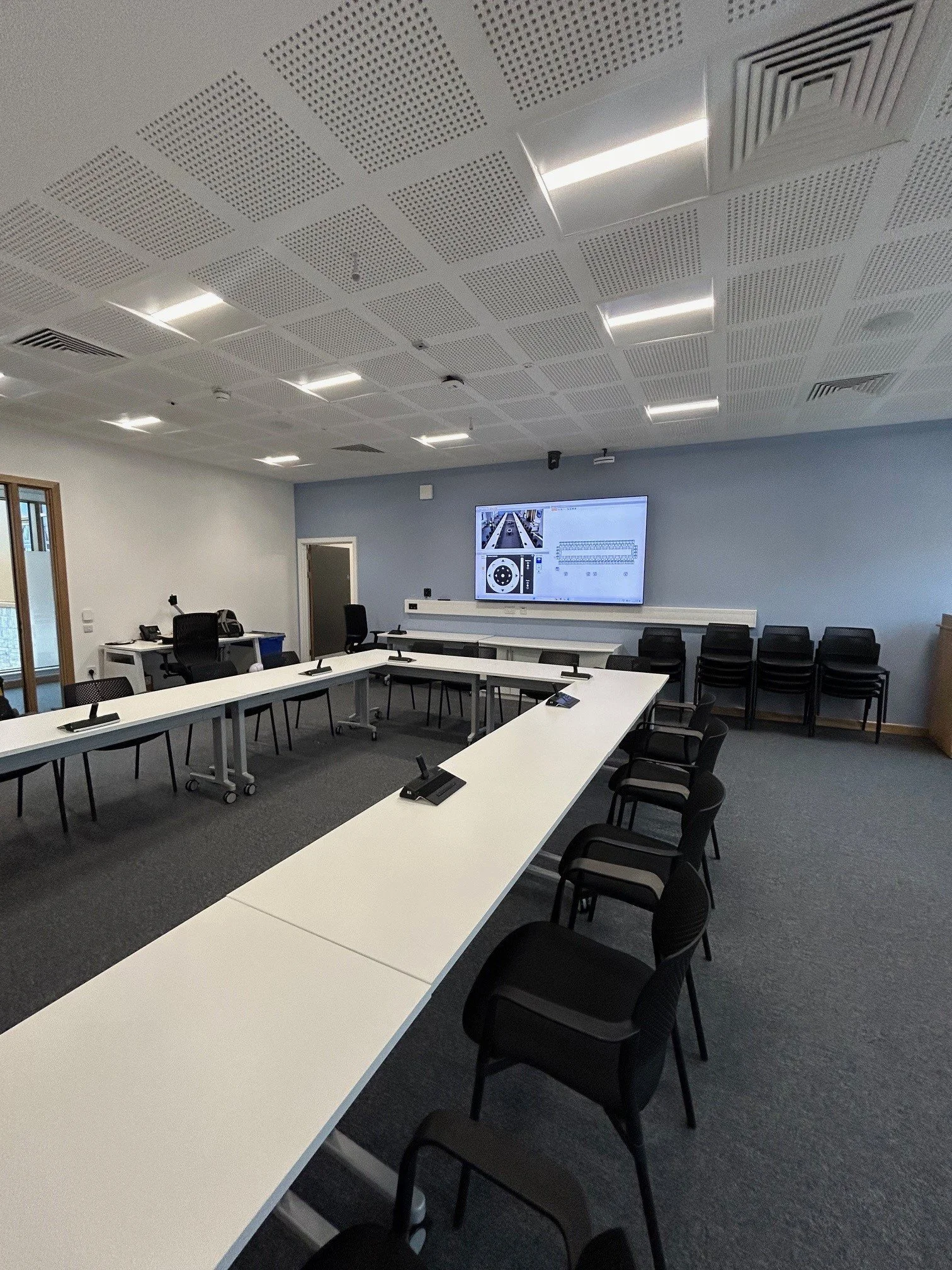 A modern conference room with a row of white tables, black chairs, a large wall-mounted screen showing presentation slides, and ceiling lights.