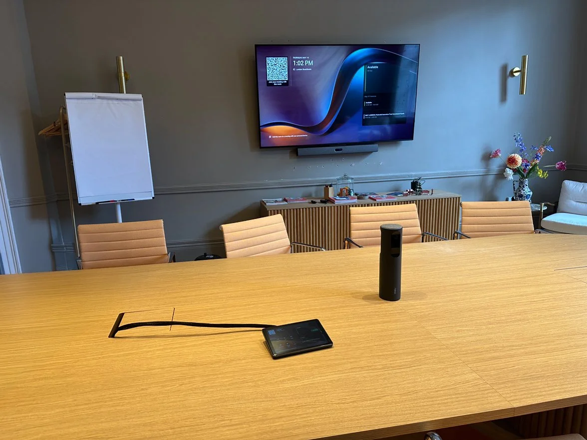 An empty conference room with a large wooden table, beige chairs, a wall-mounted TV, a whiteboard on a stand, a vase of colorful flowers, and a cylindrical speaker on the table.