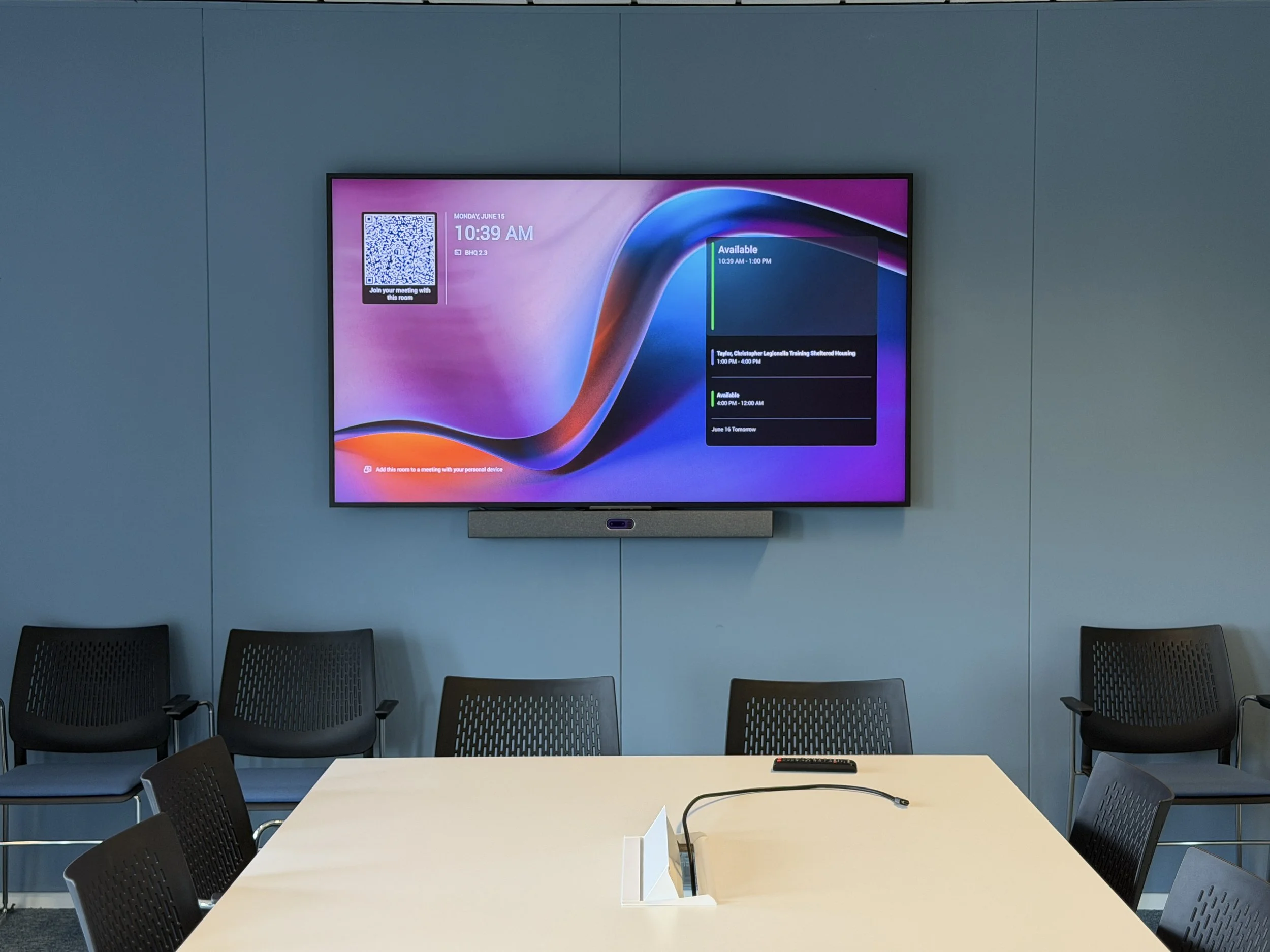 Conference room with a large screen displaying a digital schedule, surrounded by chairs, a table with a phone, and a wall with blue panels.
