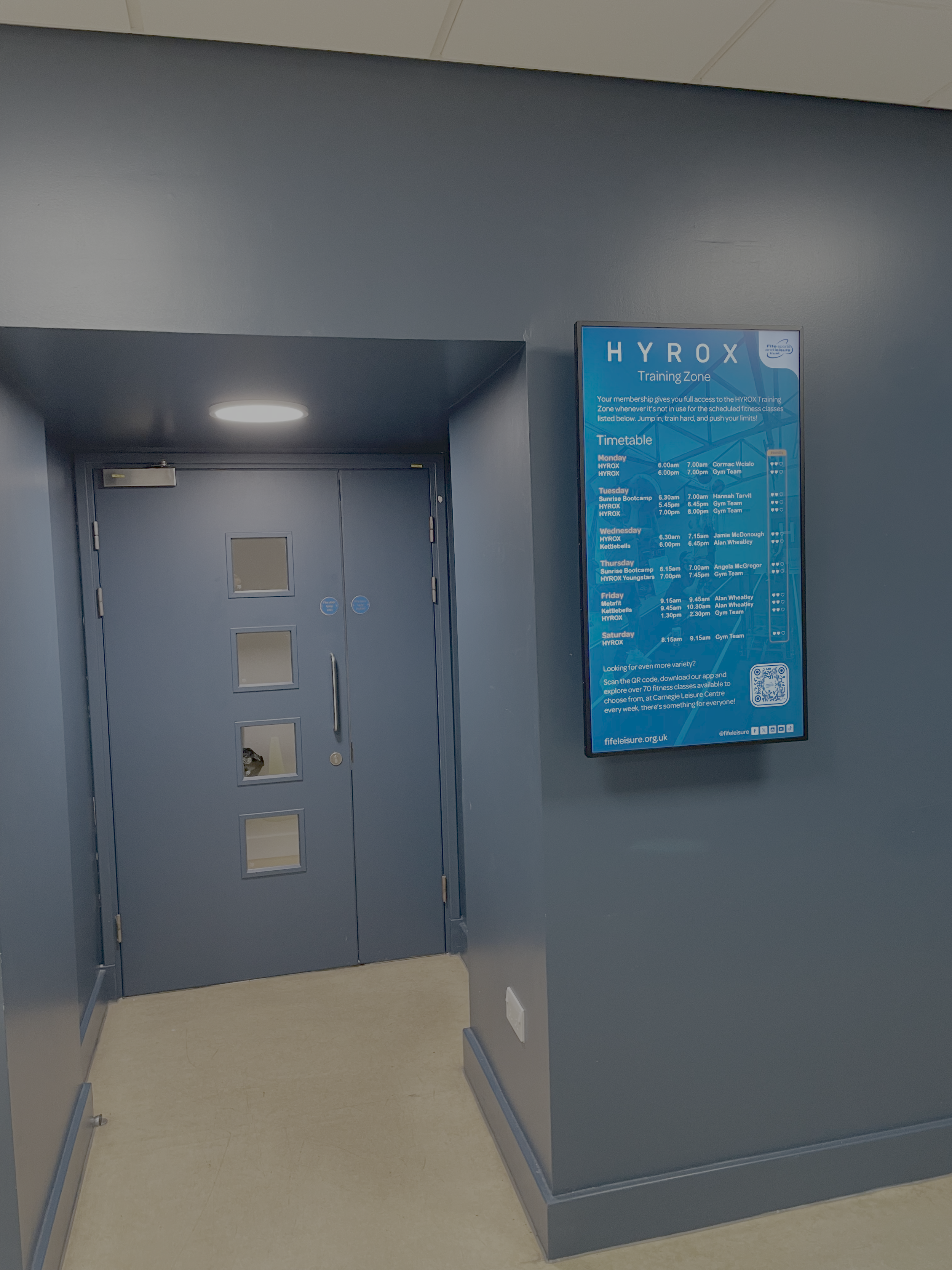 Blue hallway with a closed door and a large blue timetable sign on the wall listing fitness class schedules.