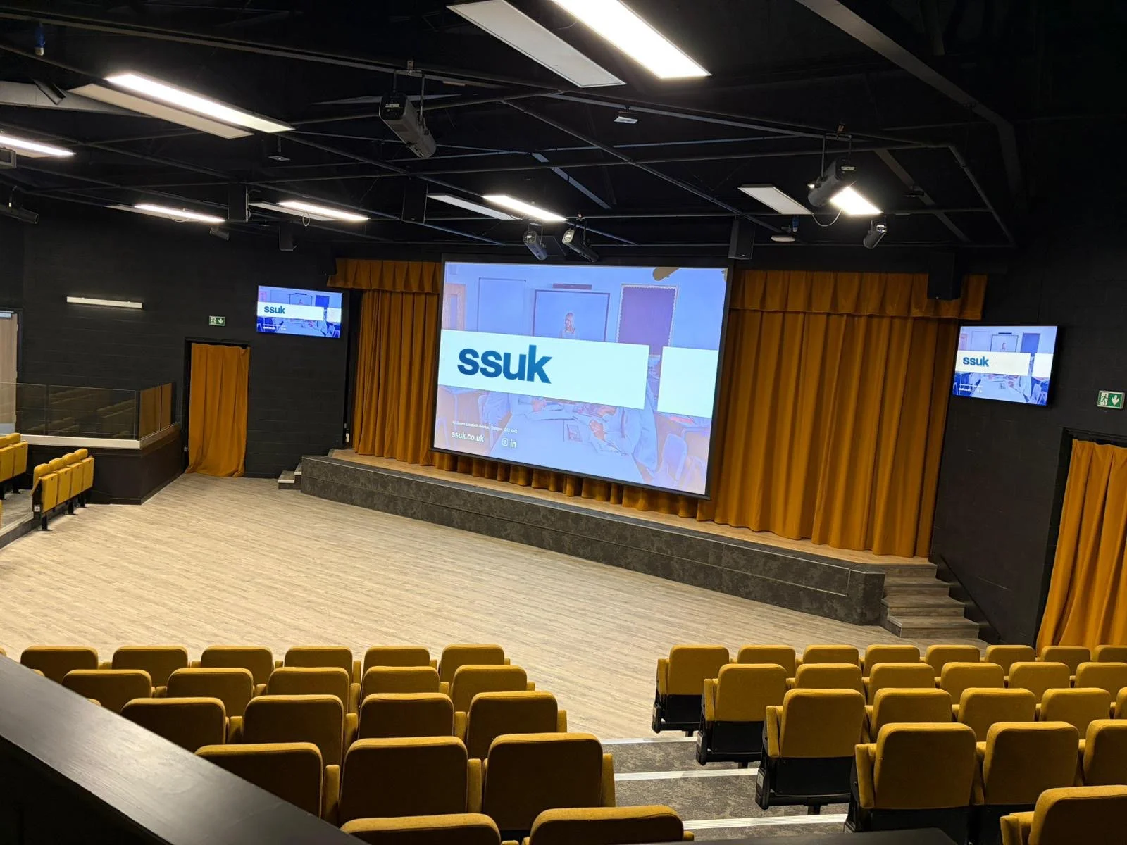 Empty auditorium with yellow cushioned seats, large stage with a projection screen displaying 'ssuk' logo, and orange curtains on the stage with black walls and ceiling.