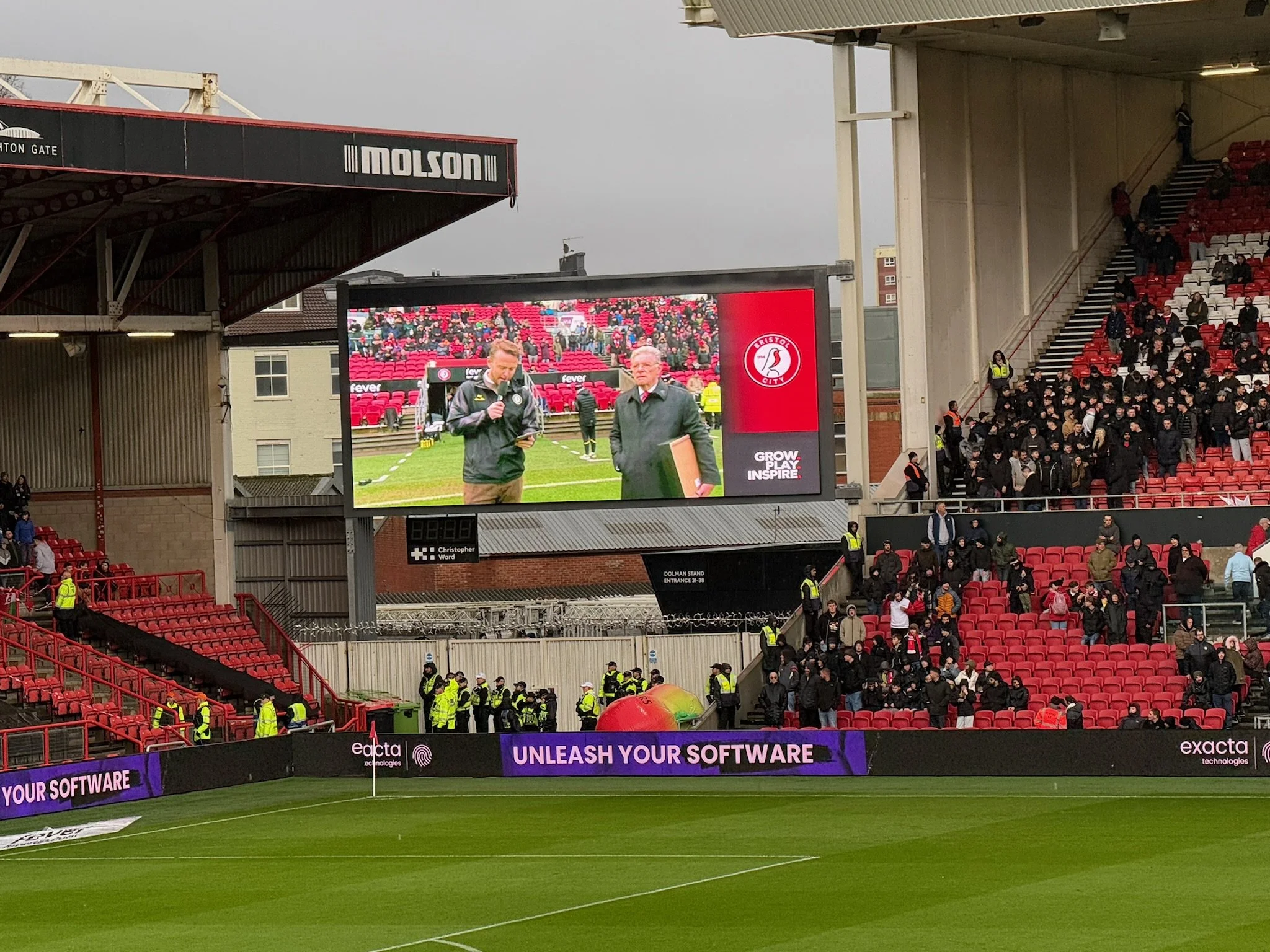 A football stadium with a large screen displaying two announcers, and spectators seated in the stands. Security personnel are visible near the field, and there are advertisements on the perimeter.