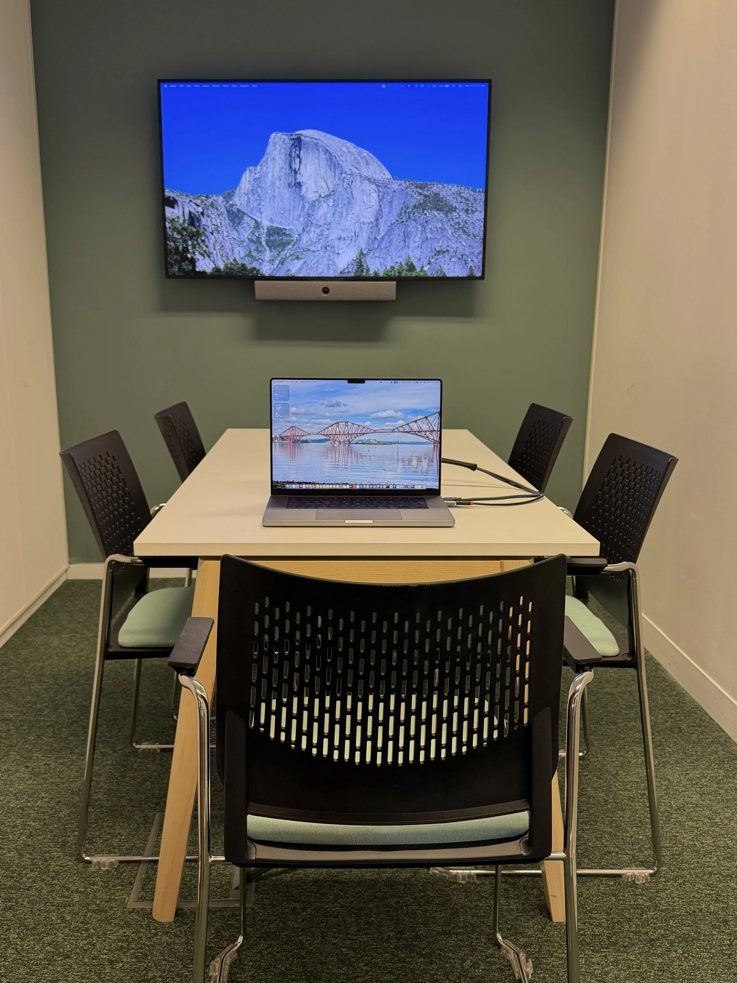 A conference room featuring a white table with a MacBook laptop, and a large mounted flat-screen monitor displaying a mountain landscape with a bright blue sky.
