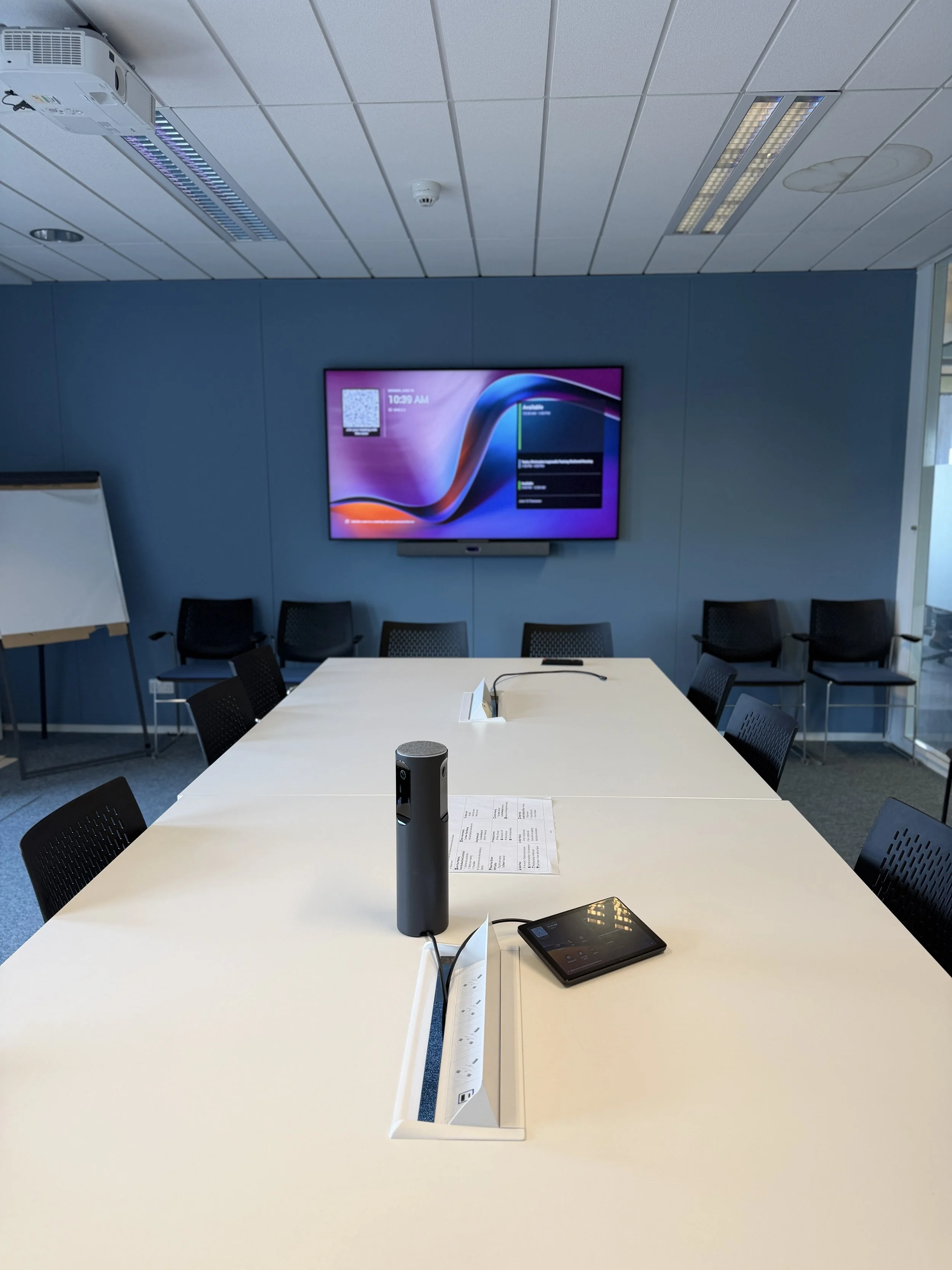 Conference room with a large white table, chairs, a wall-mounted flat screen TV, and electronic devices on the table, including a cylindrical speaker.
