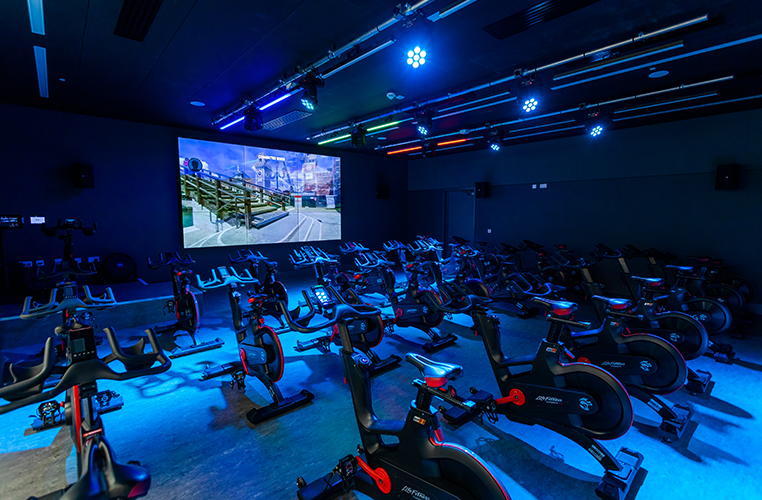 Indoor spin class room with multiple exercise bikes illuminated by blue and green lights, large screen at the front displaying a scenic outdoor cycling route.