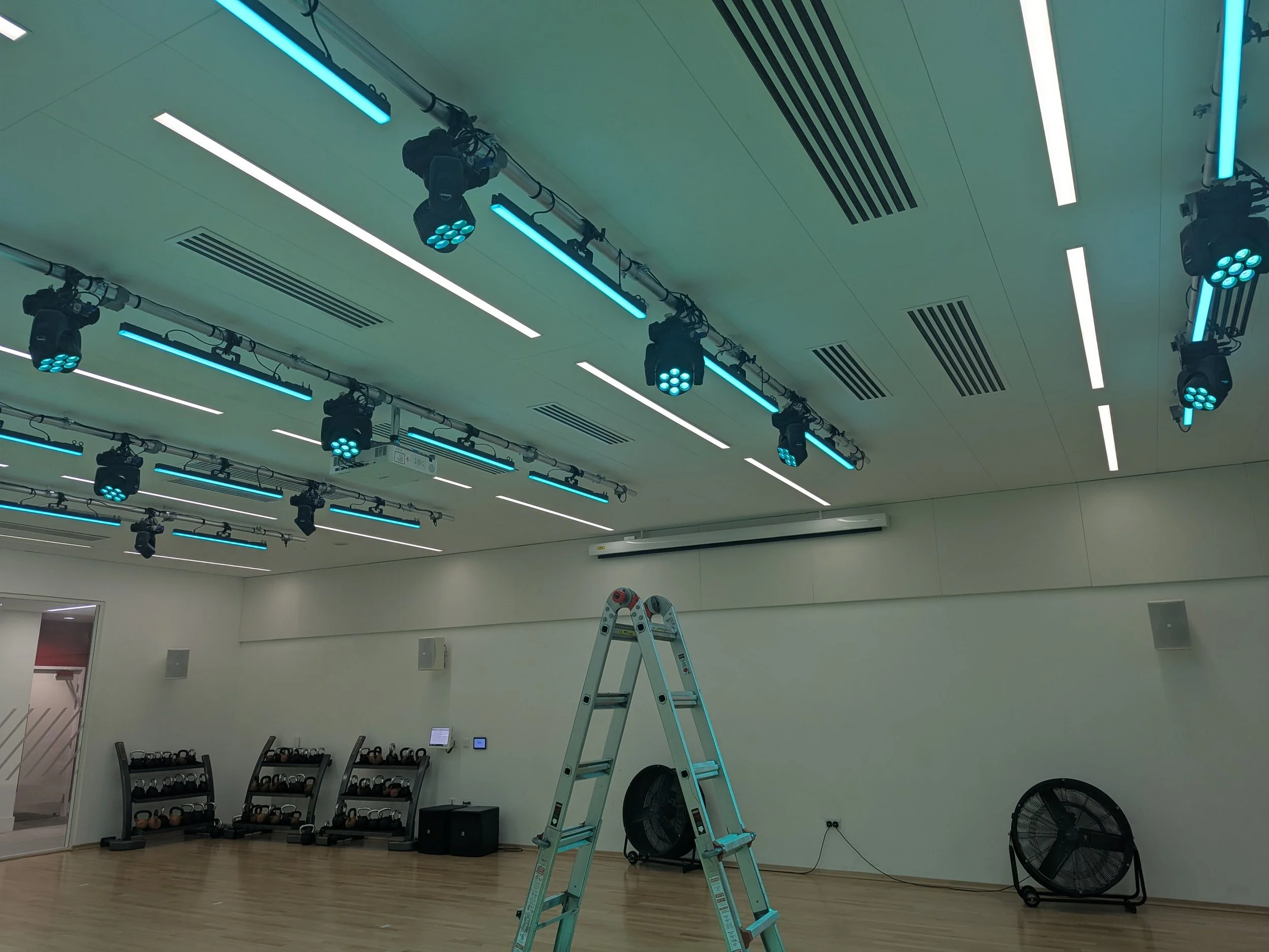 An empty gym room with a ladder, black fans, weight racks, and blue electric light bars on the ceiling.