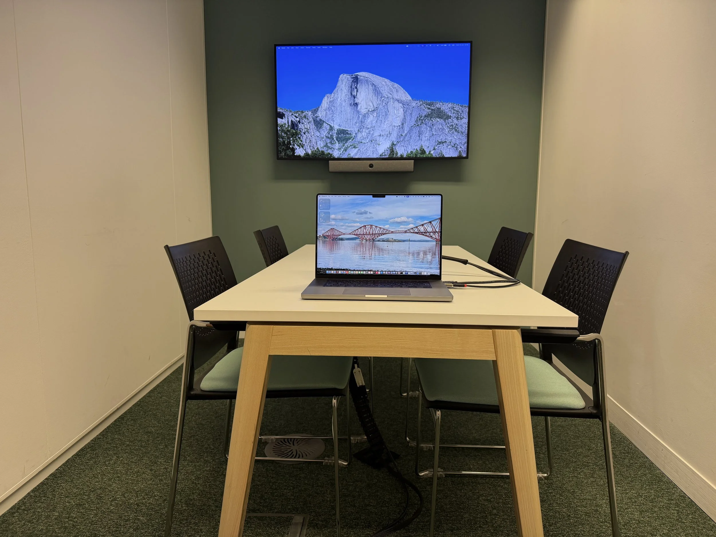 A meeting room with a white table, four black chairs and a computer monitor displaying a bridge over water.