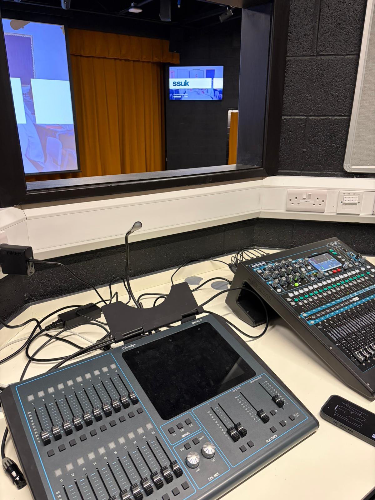 Inside a soundproof recording booth, a mixing console and audio equipment are placed on a white table. The window shows another room with a wooden door and a monitor displaying the ssuk logo.