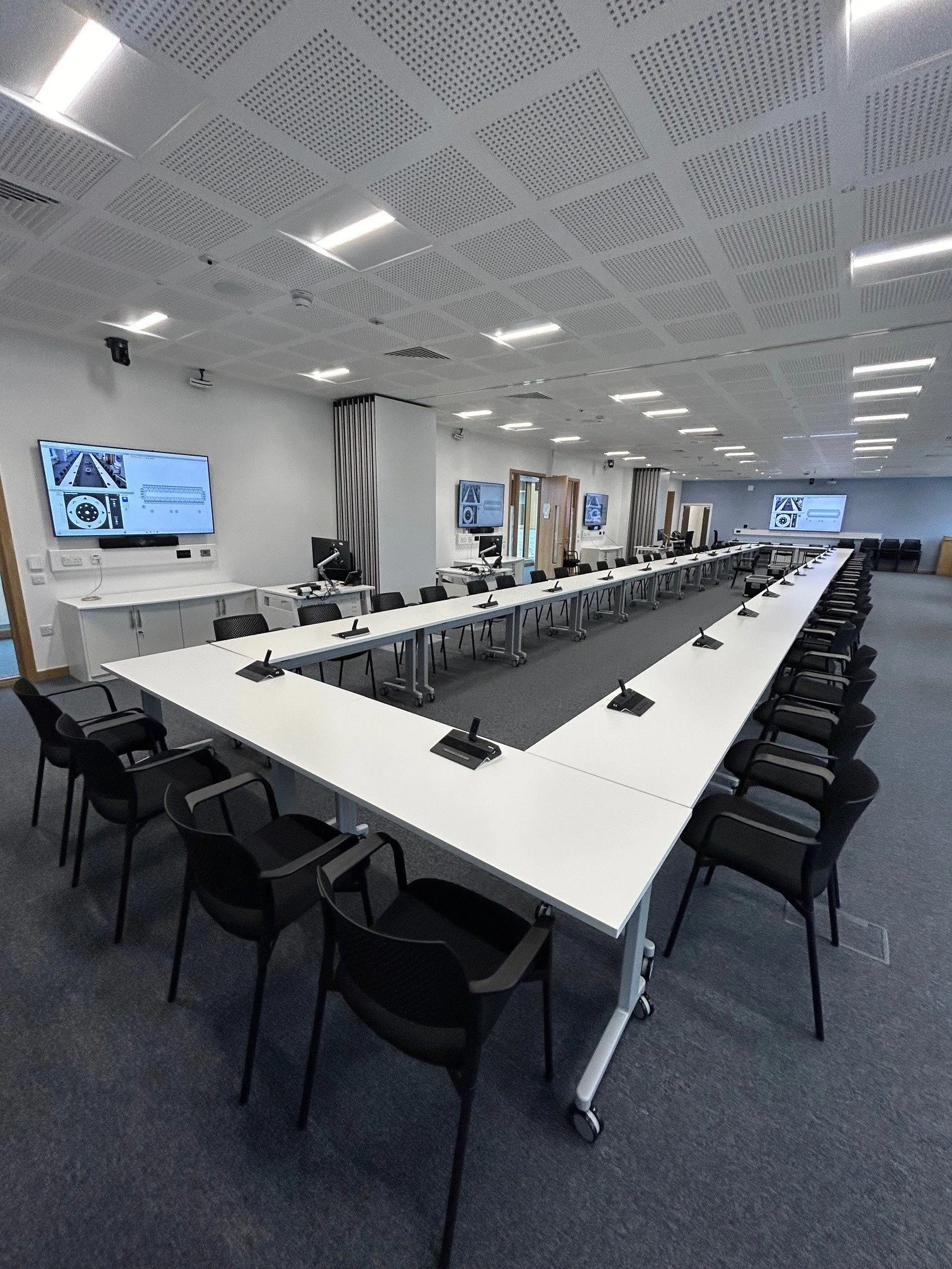 A modern conference room with a U-shaped arrangement of white tables, black chairs, computer monitors, and microphones. Multiple large screens display technical diagrams.