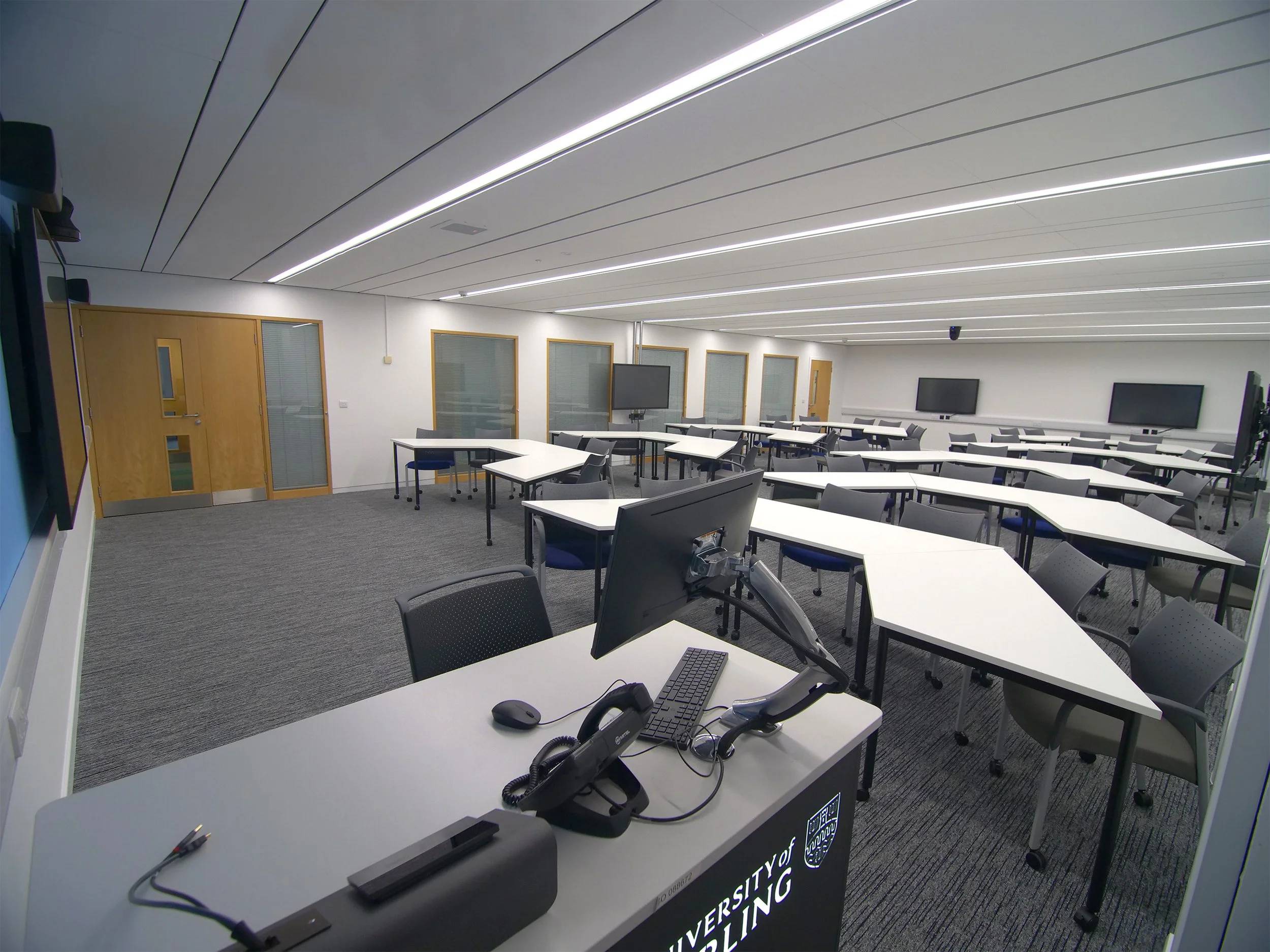 A modern conference room with white tables, black chairs, and multiple flat-screen TVs mounted on the white walls. The ceiling has long, white, linear lighting fixtures. There are closed wooden doors with glass panels along one wall, and computer monitors and conference equipment on the tables.