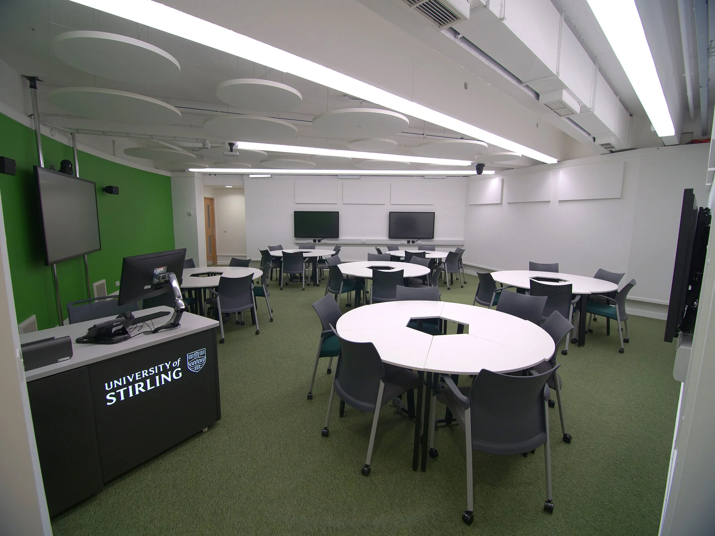 A modern classroom or conference room with white round tables and black chairs, two large wall-mounted screens, green and white walls, and a ceiling with circular acoustic panels and fluorescent lights.
