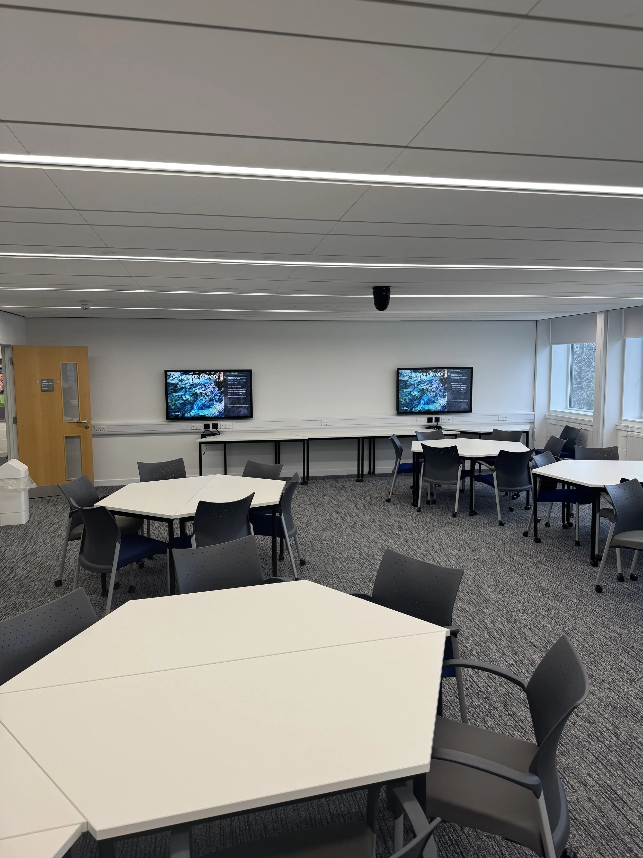 A meeting room with white tables and gray chairs, two wall-mounted screens displaying graphics, and a door on the left side of the room.