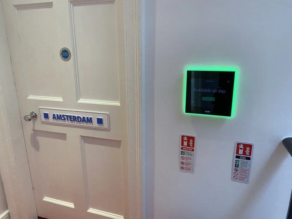 A white door with a blue and white sign that is next to a digital screen with a green light border on a white wall.