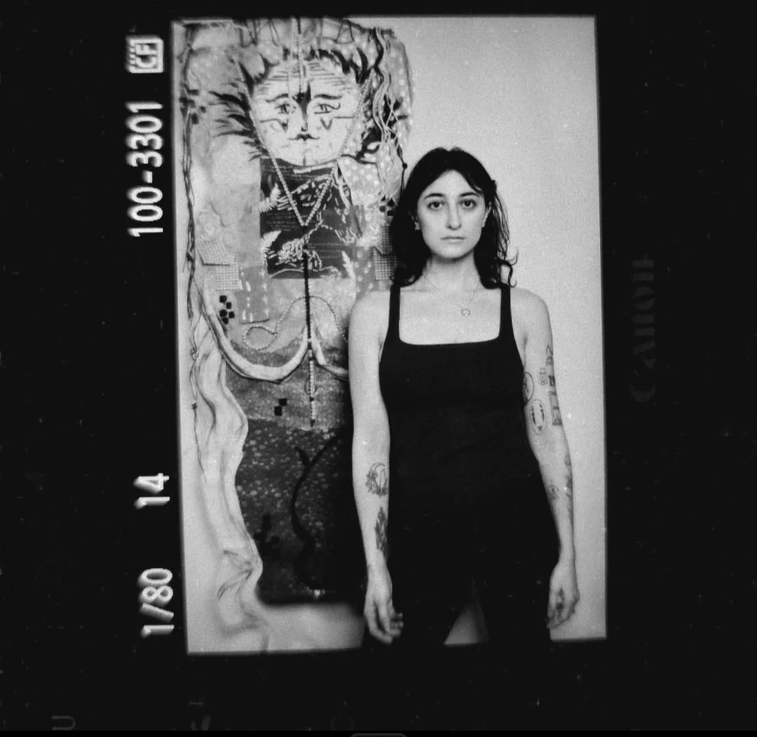 Black and white photograph of a young woman with tattoos standing against a painting of a face on the wall.