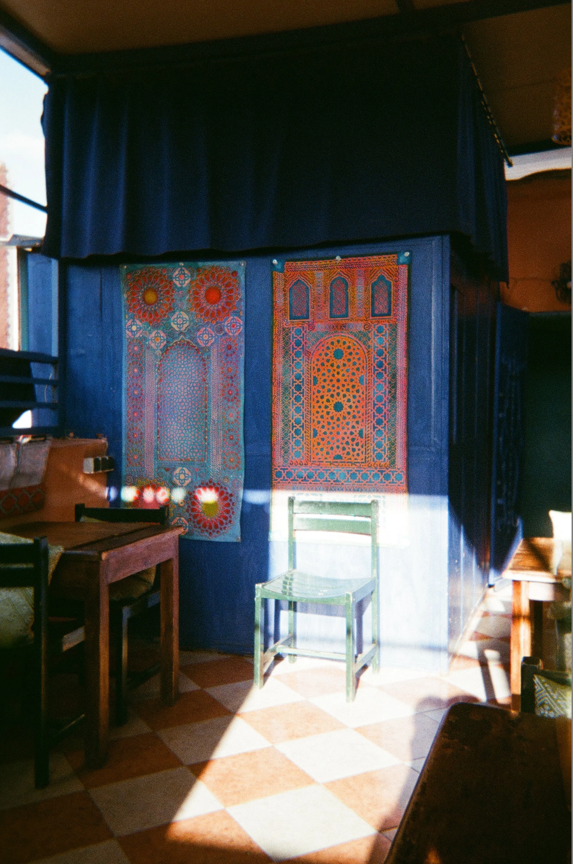 Interior of a room with blue walls and colorful decorative wall hangings, a green transparent chair, wooden tables and chairs, and patterned tiled floor with sunlight casting shadows.