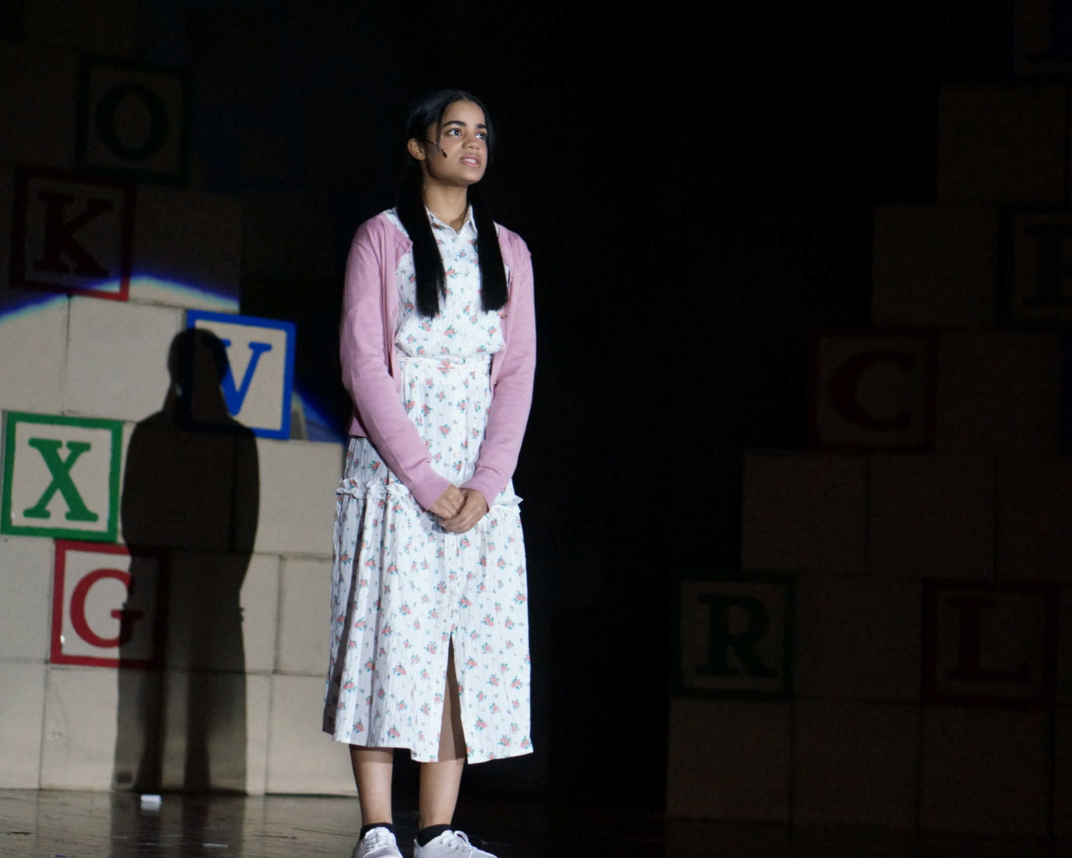 Young woman with long dark hair, standing on stage in a dark room, wearing a white floral dress with a pink cardigan and white sneakers.