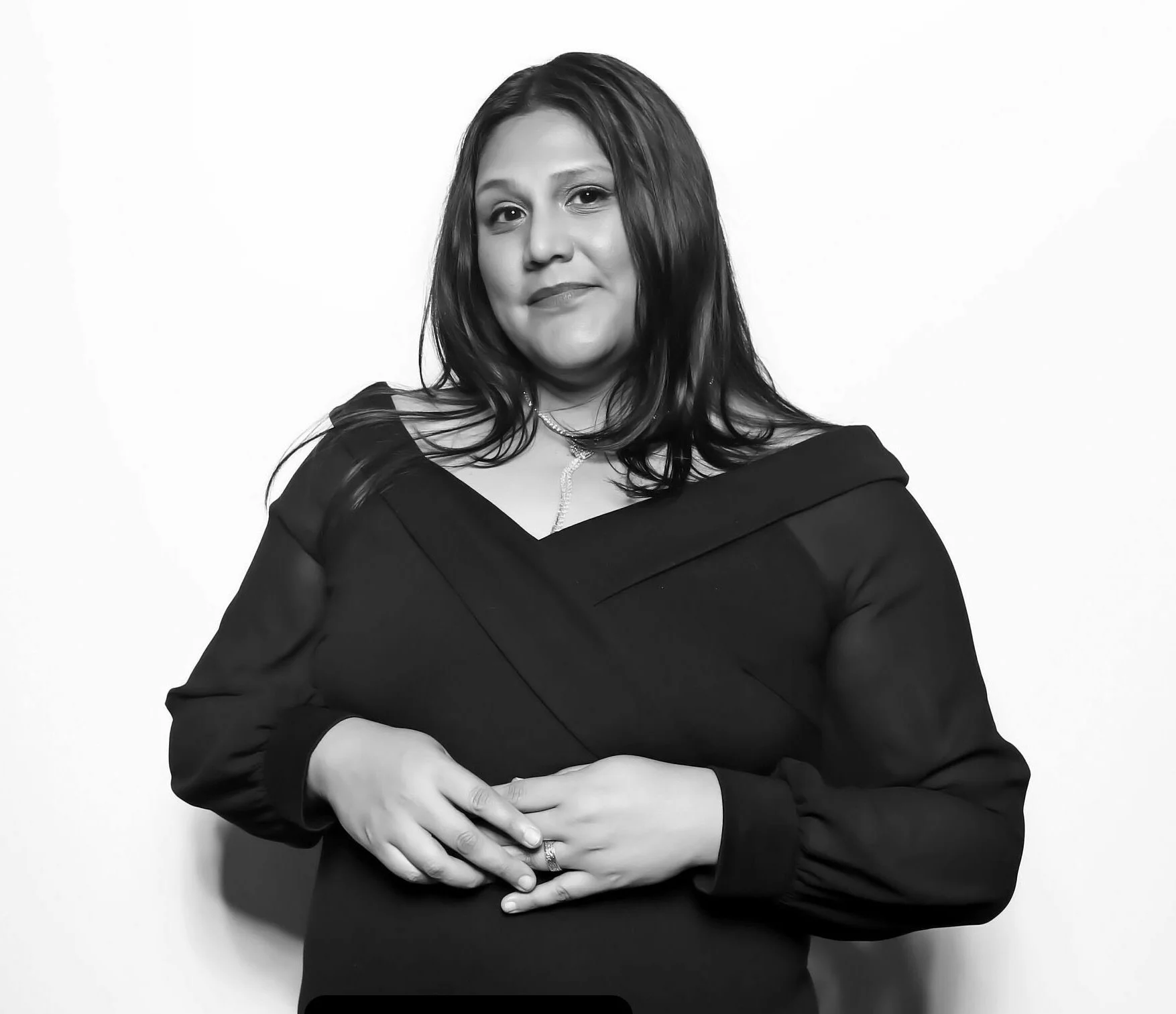 Black and white portrait of a woman with long dark hair wearing a dark dress with an off-the-shoulder neckline, standing against a plain light background, with her hands gently clasped.