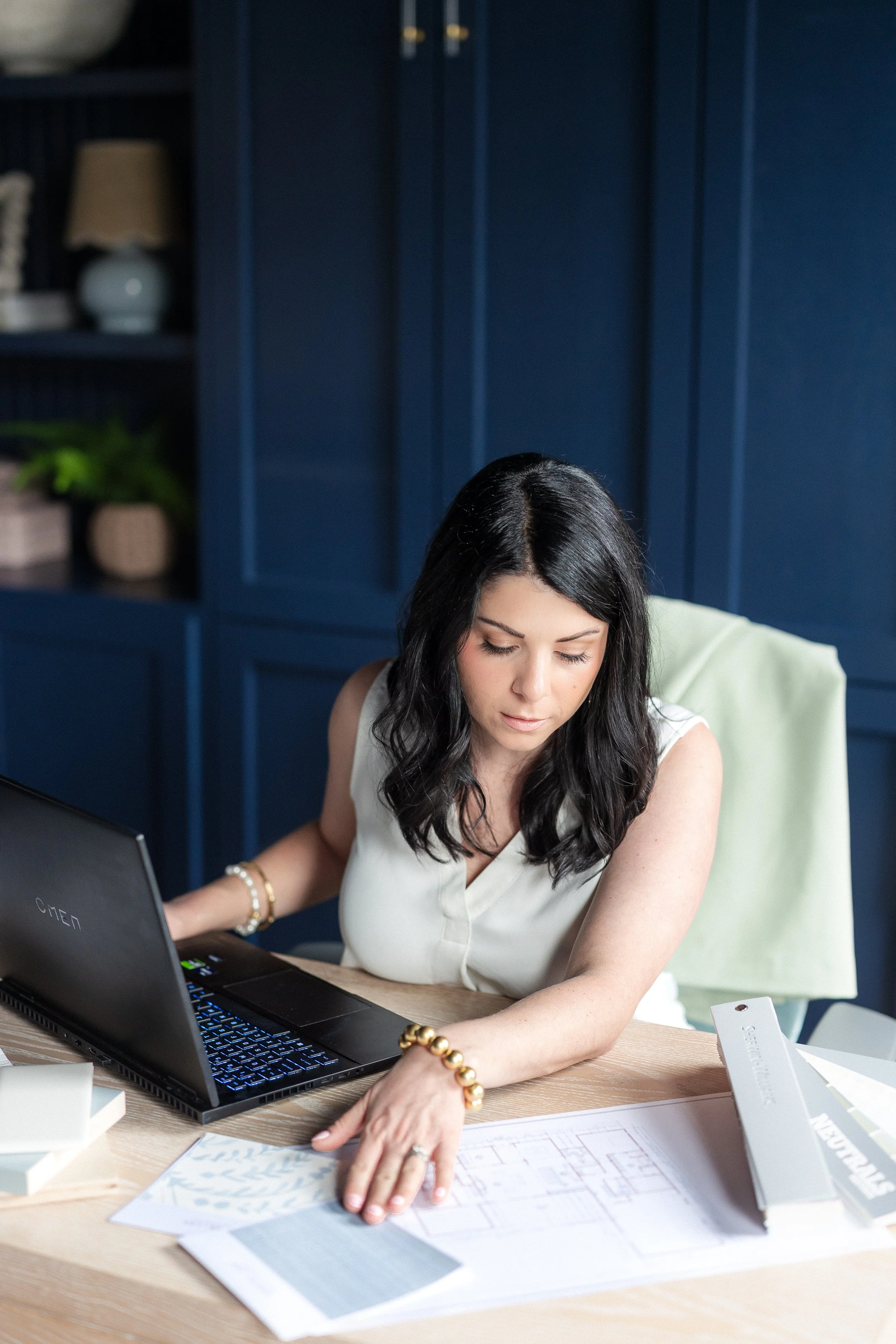 Woman sitting at desk with laptop and blueprints, in a room with navy blue cabinets and decor.