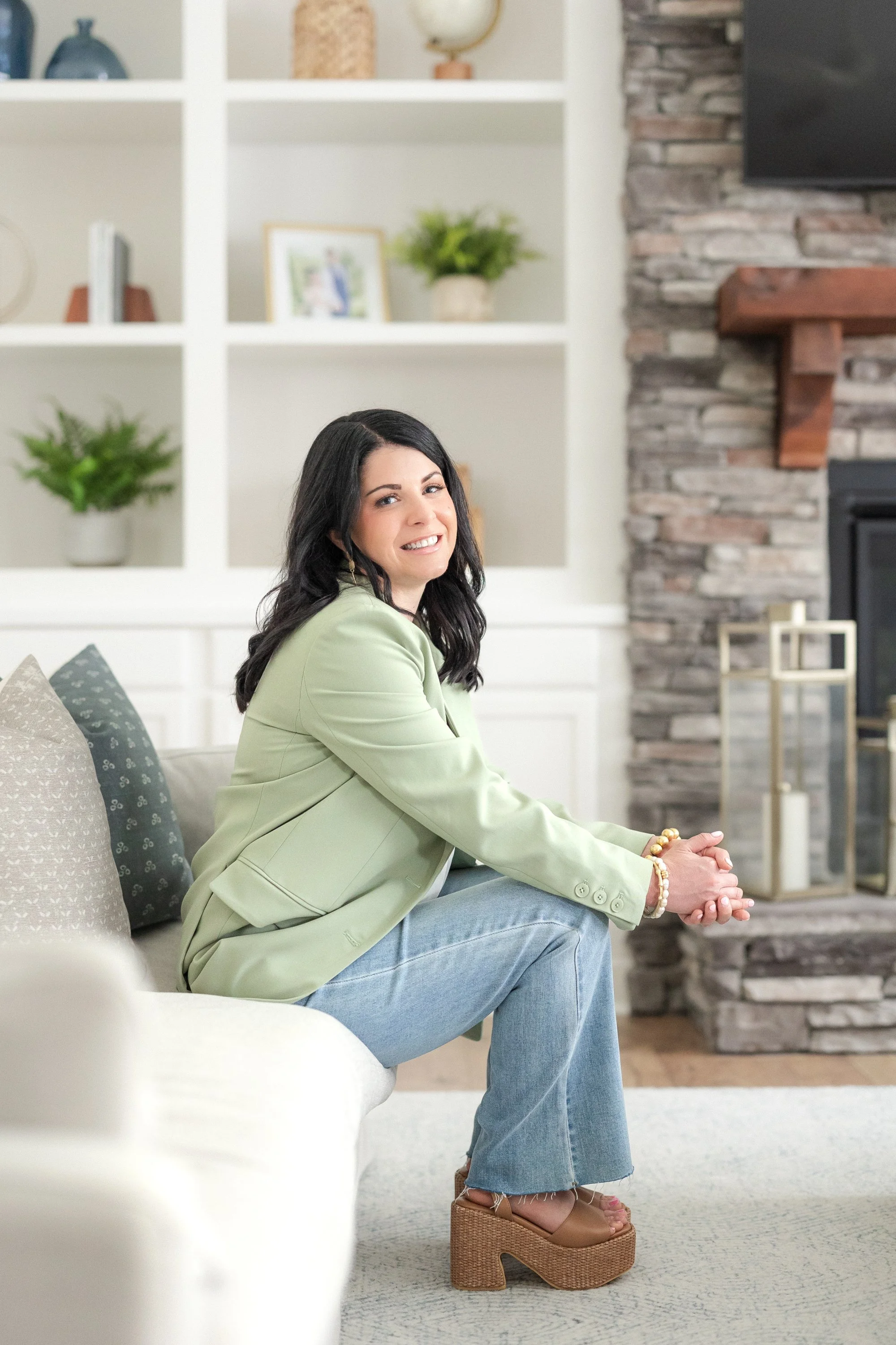 A woman with black hair dressed in a light green blazer, blue jeans, and platform sandals sitting on a white sofa in a living room with a stone fireplace and decorative shelves.