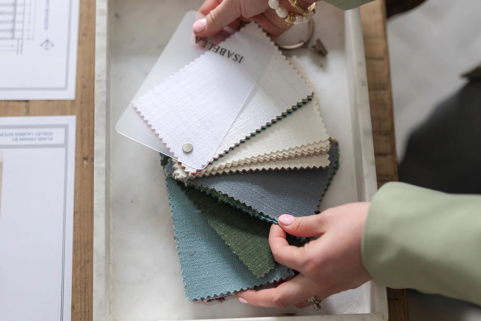 Person holding fabric swatches and a sample card with fabric textures and colors in shades of white, gray, green, and blue, on a white surface.