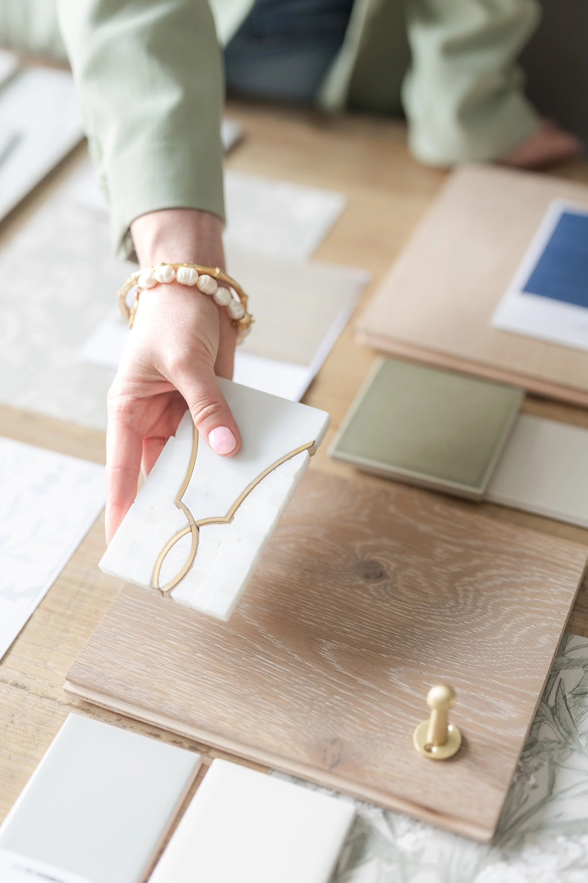 A person holding a white marble tile with gold accents above a wooden surface, with various tile samples and color swatches spread out on the table.