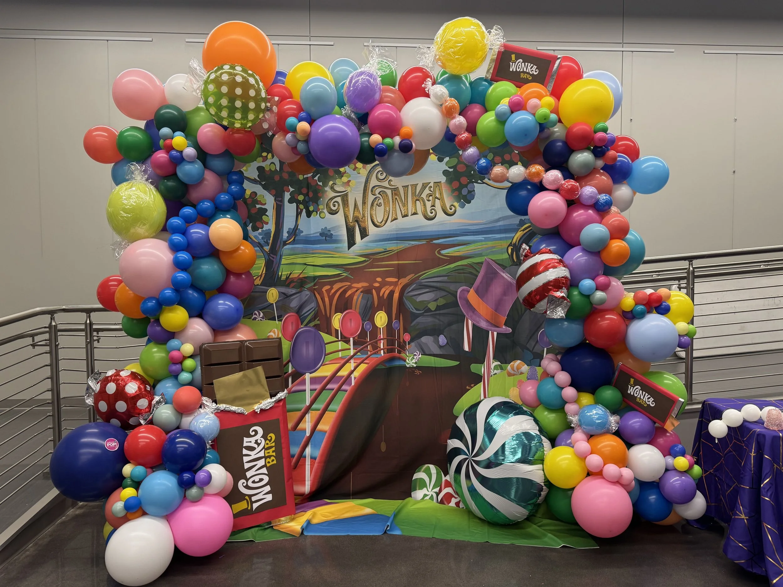 A colorful balloon display featuring various sizes and colors, including pink, blue, green, orange, purple, red, and white balloons, arranged around a background with 'Wonka' branding and candy-themed decorations at an event or celebration.