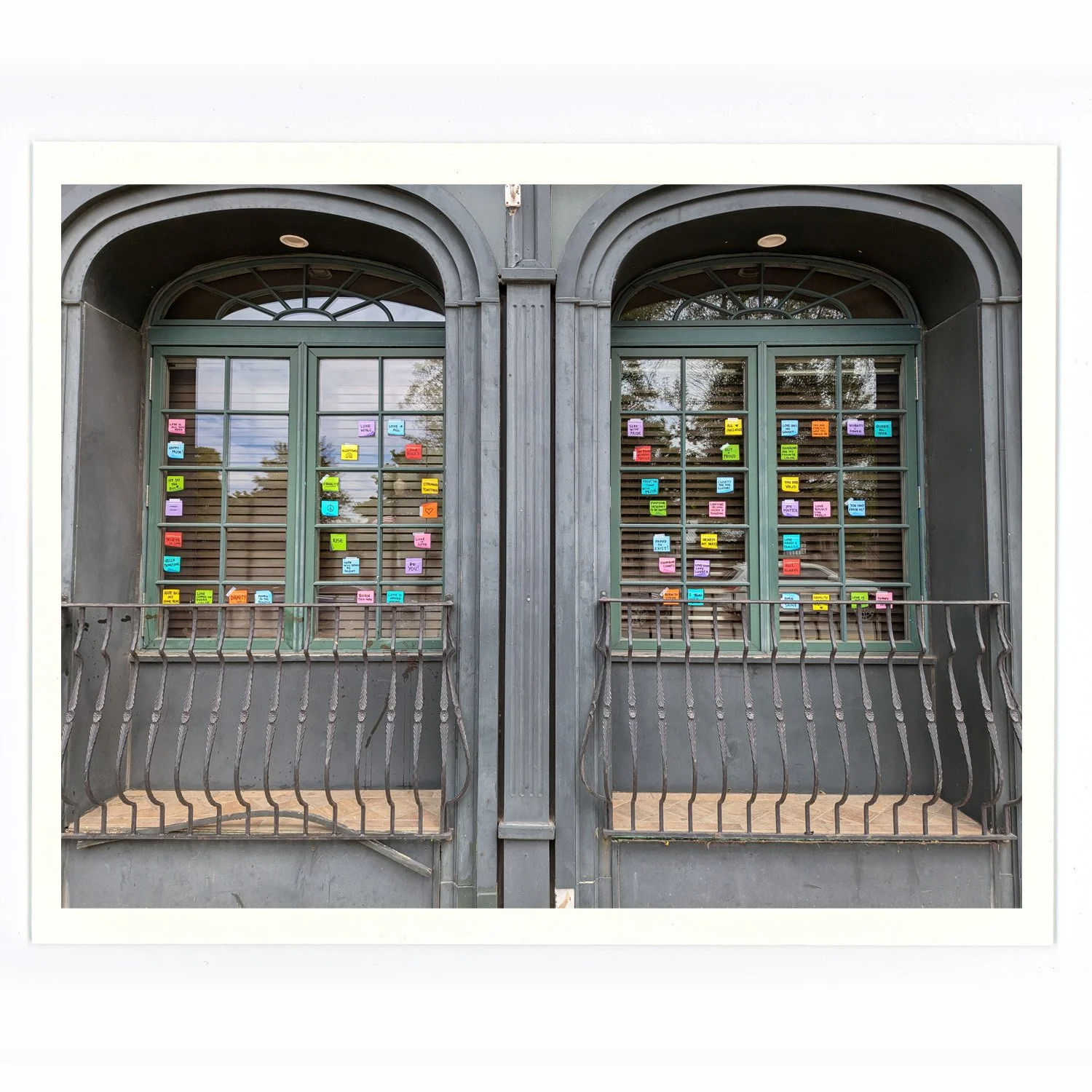A pair of green framed windows with stickers on the glass, set in a gray building with arched openings and a black metal railing in front.