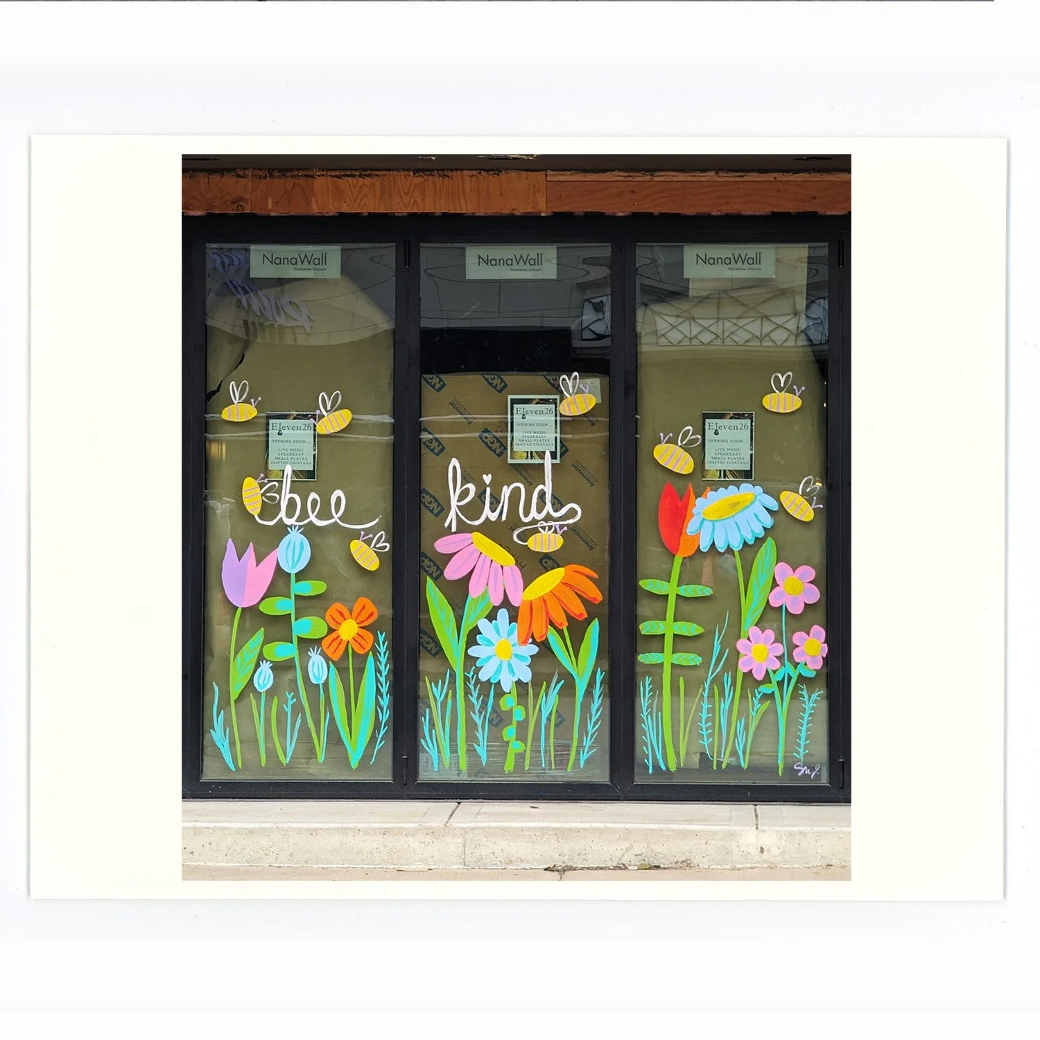 Decorated window with colorful paper flowers and bees, displaying the words 'be kind' in white script.
