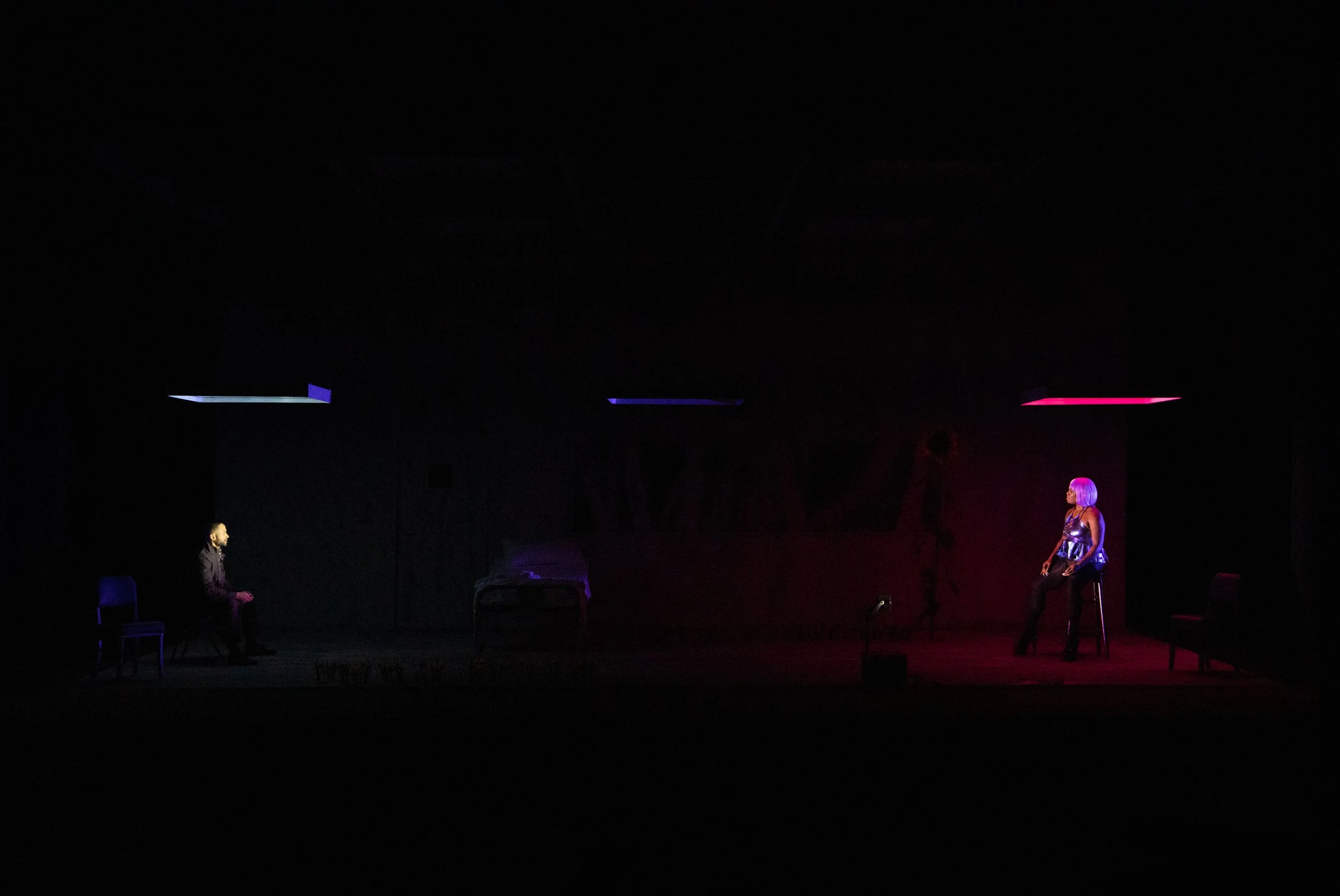 A dimly lit stage scene with two individuals sitting apart under spotlights; one under blue light on the left and the other under pink light on the right. A bed is in the center.