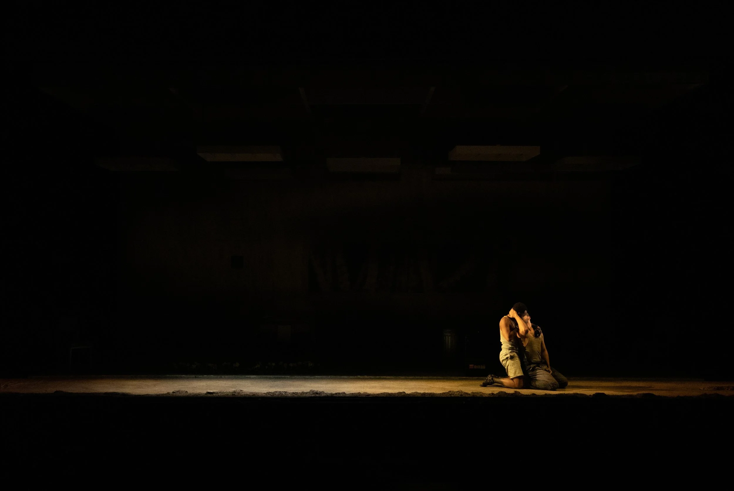 Two performers embracing on a dimly lit stage, with one kneeling and the other sitting.